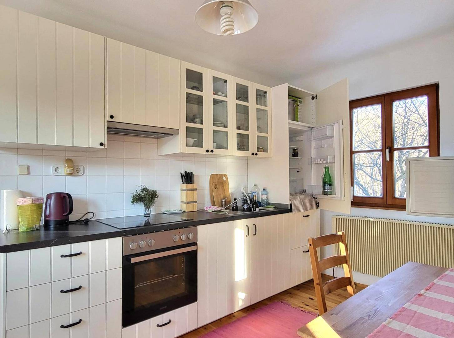 Helle Küche mit weißen Schränken, schwarzer Arbeitsplatte, Backofen und Esstisch am Fenster.