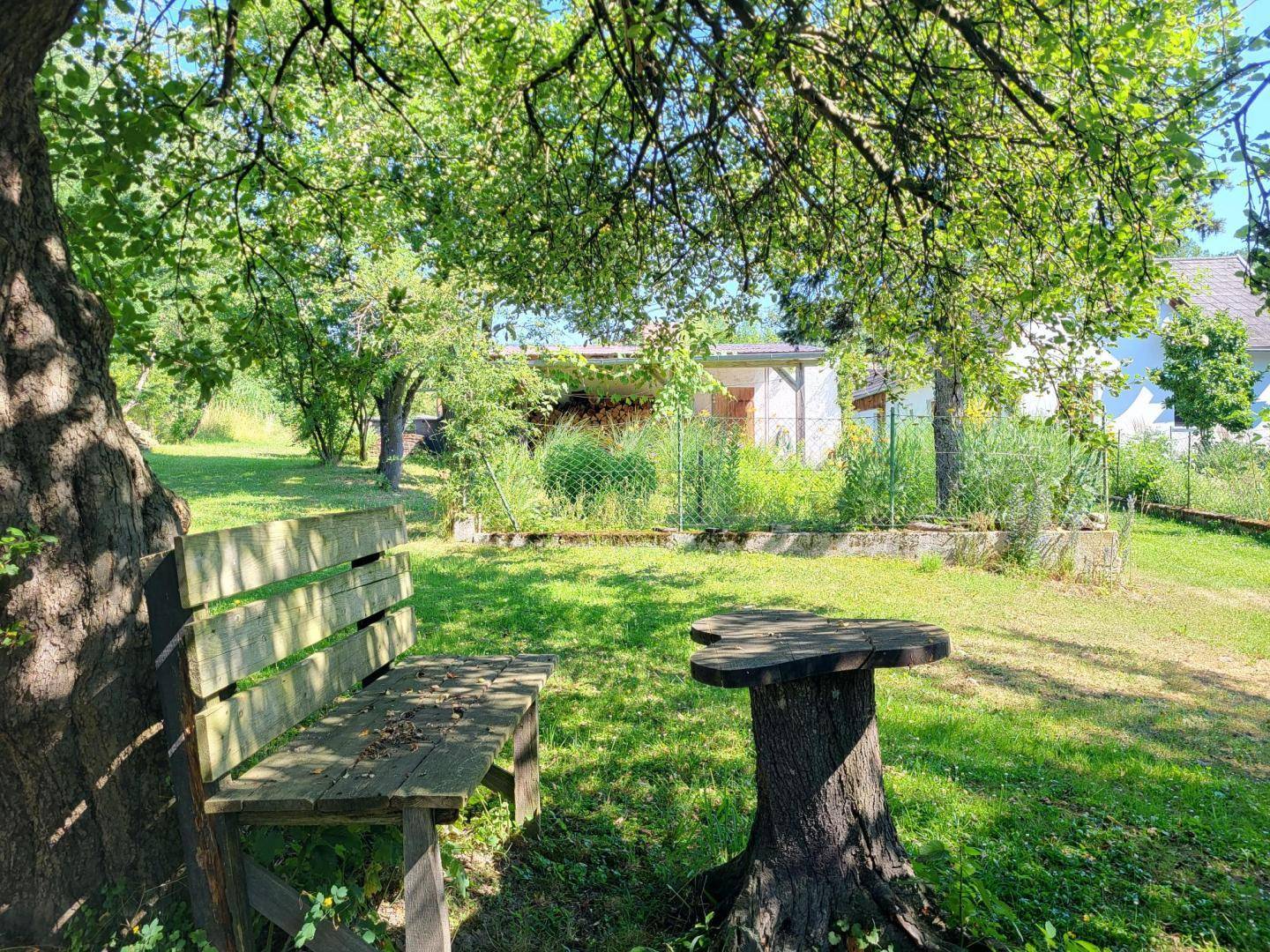 Idyllischer Gartenbereich mit einer Holzbank und einem Baumstamm-Tisch unter schattenspendenden Bäumen.