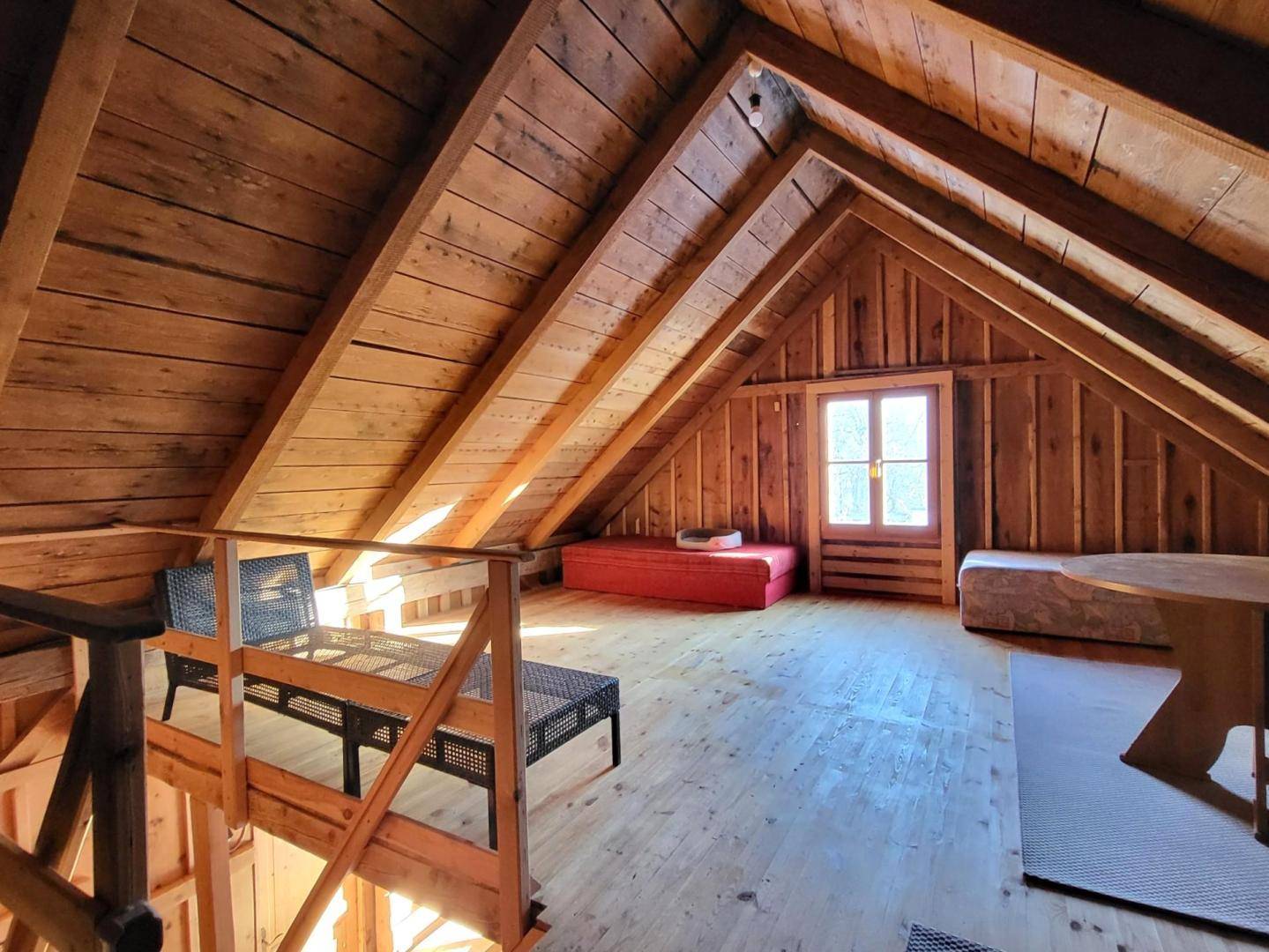 Helles Dachgeschoss mit sichtbaren Holzbalken, Holzboden und einem Fenster für Tageslicht.