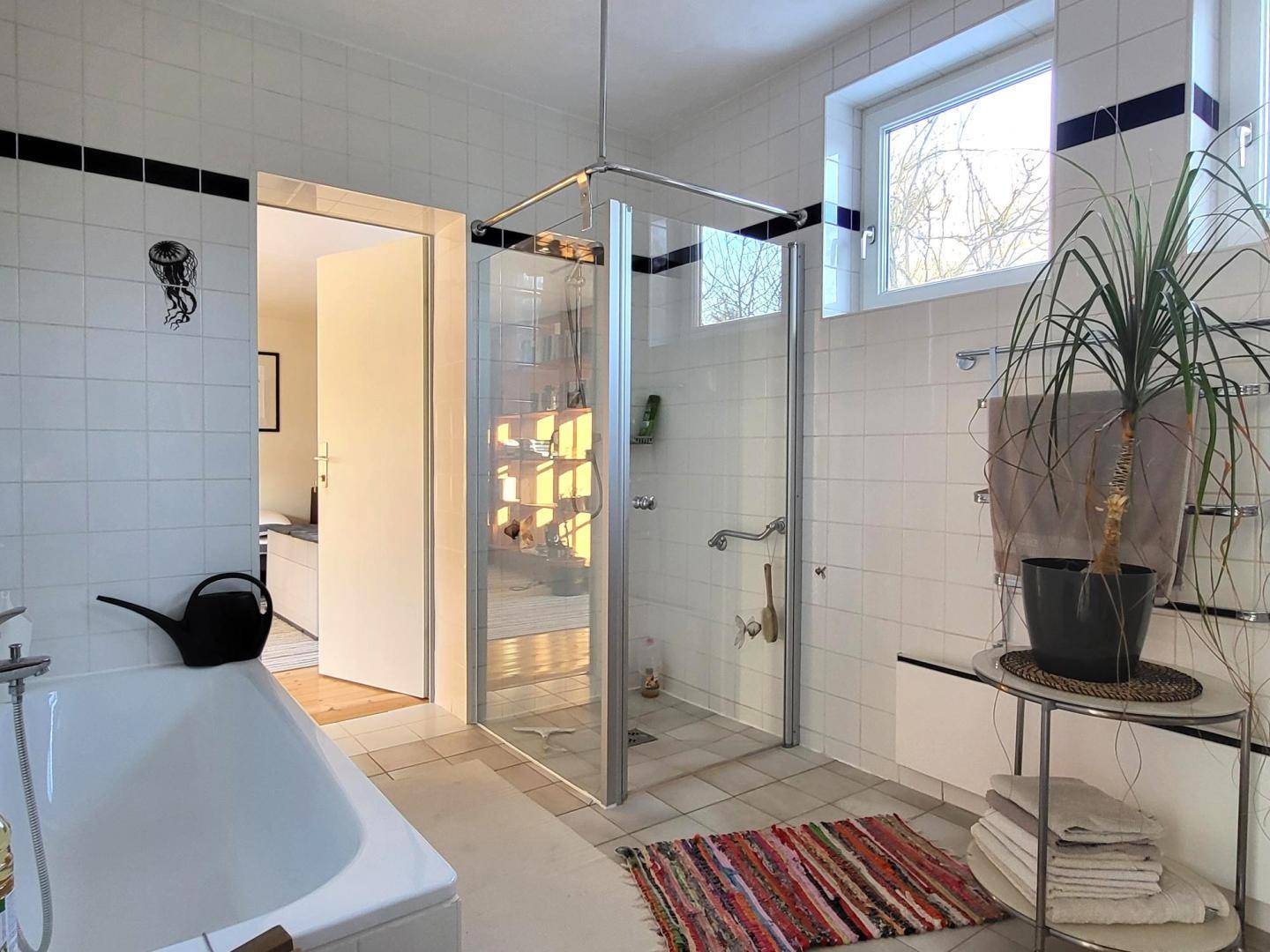 Helles Badezimmer mit Badewanne, Duschkabine, weißen Fliesen und einem Fenster für Tageslicht.