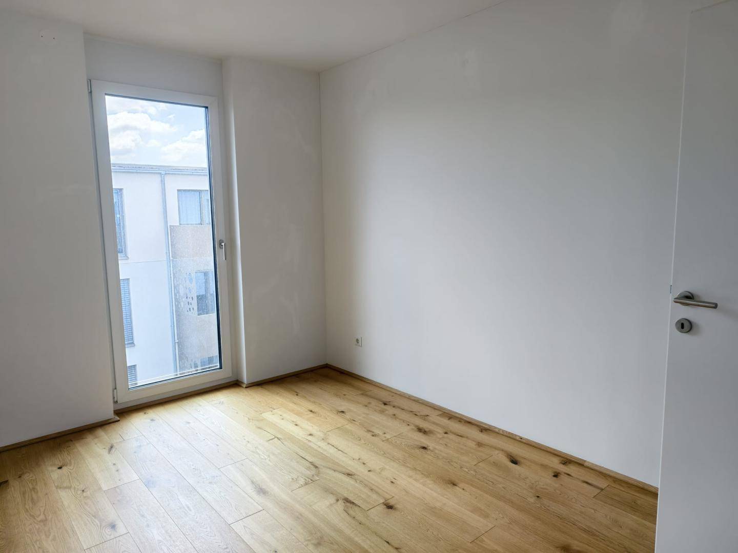 Helles Zimmer mit Holzboden und einer hohen Fenstertür, die viel Licht hereinlässt.