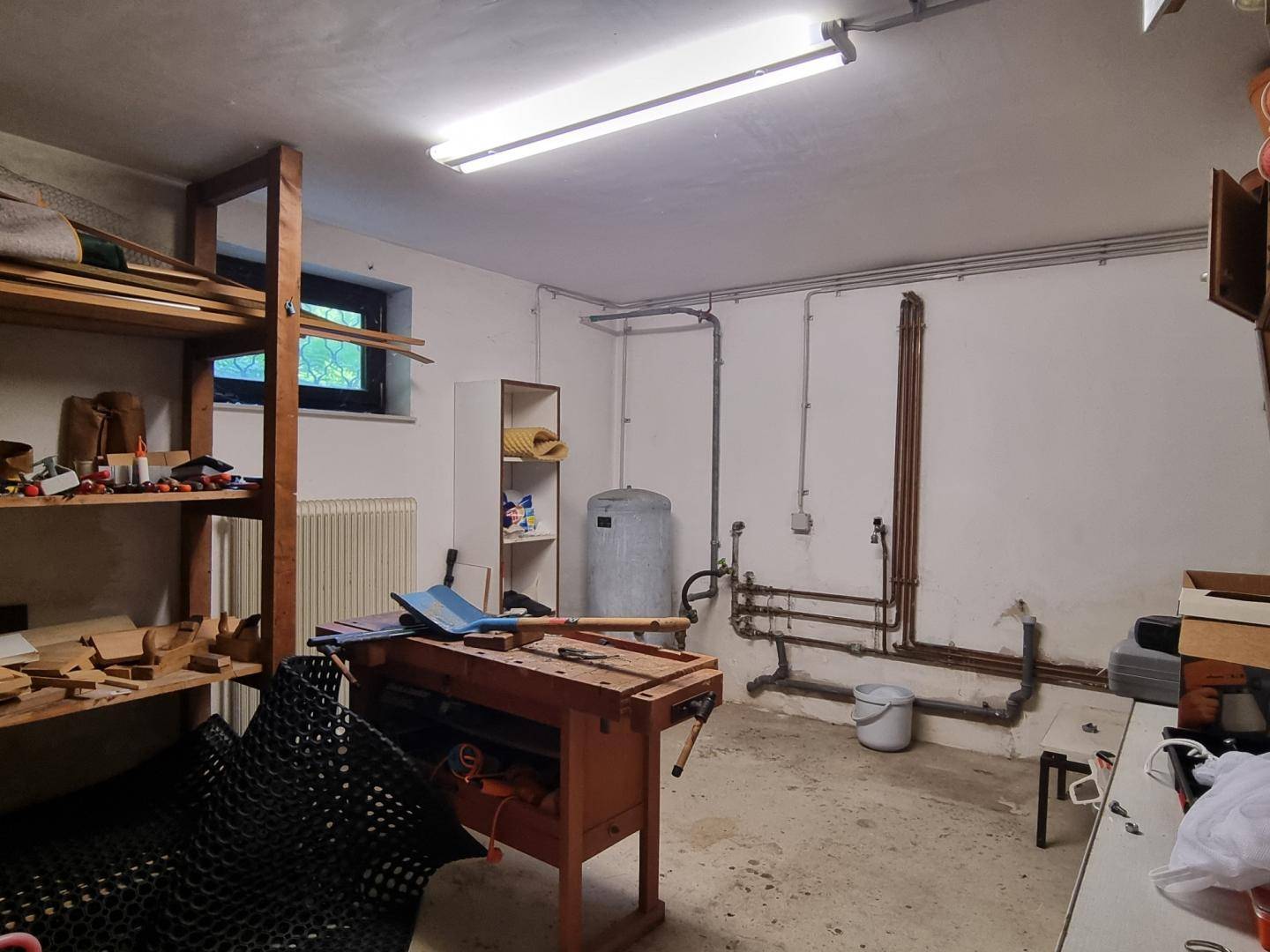 Werkstatt im Keller mit Werkbank, Regalen und sichtbaren Installationen.