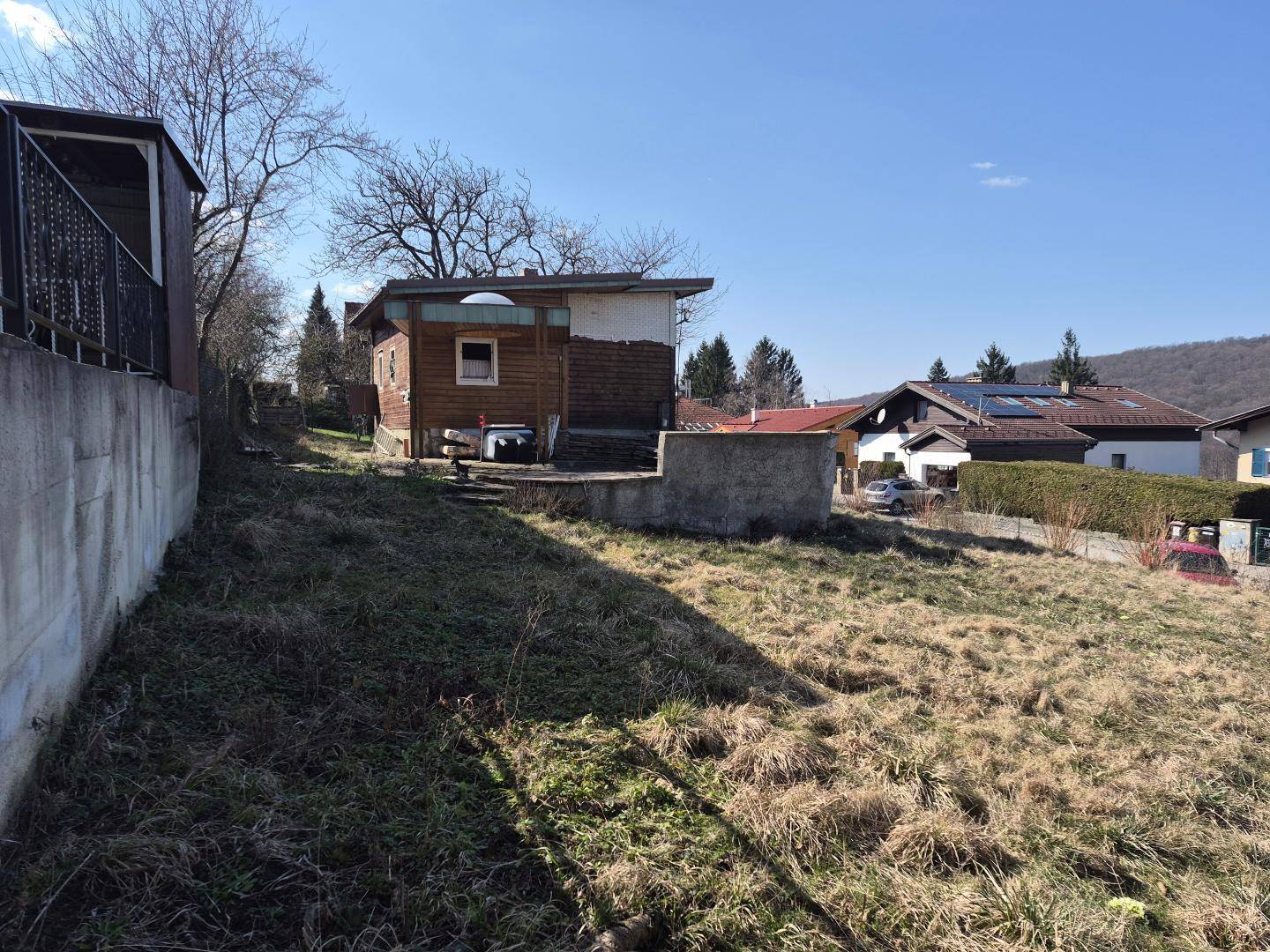 Kleines Holzgebäude neben einer Betonmauer, umgeben von trockenem Gras unter klarem Himmel.