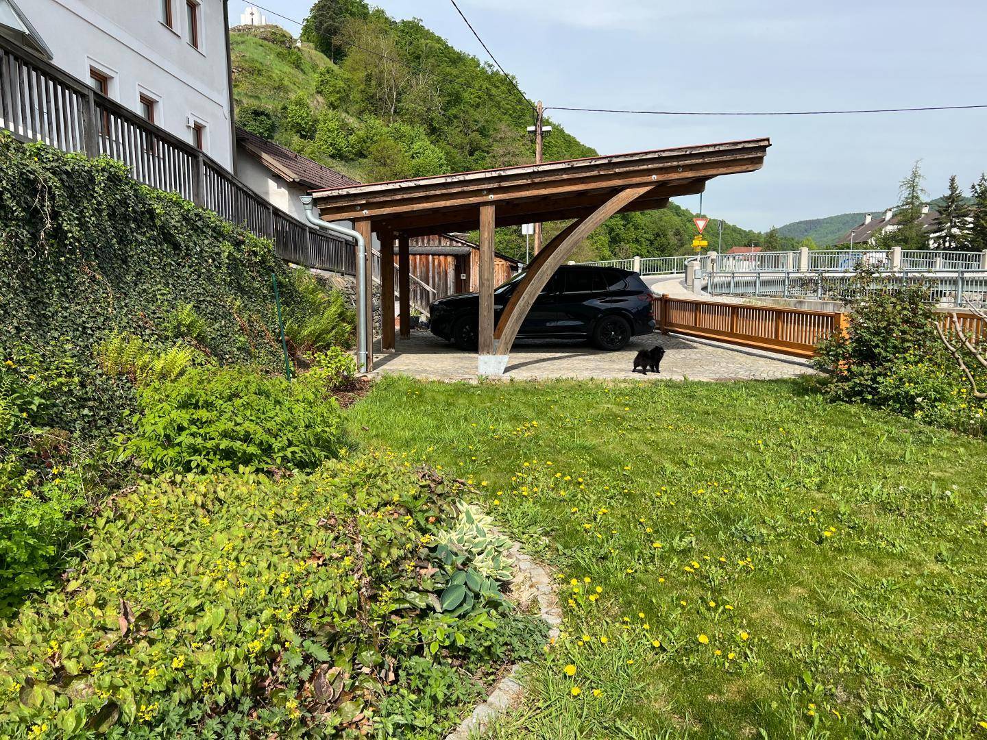 Grüner Gartenbereich mit Rasen, Sträuchern und einem Holzcarport für ein Fahrzeug.