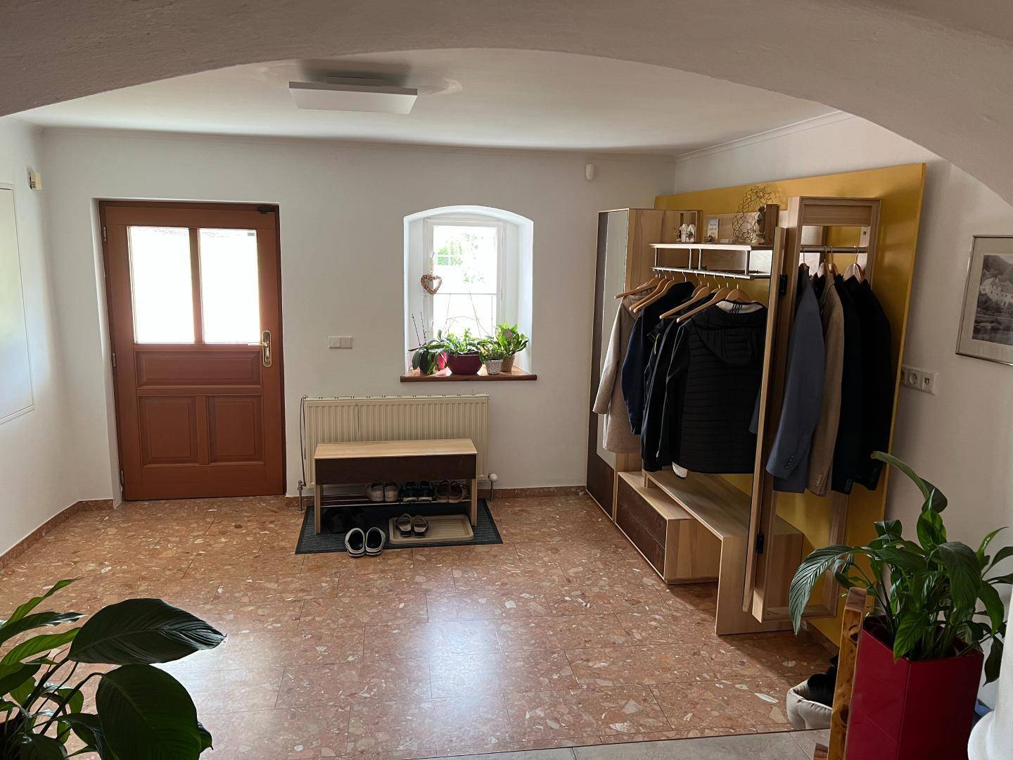 Eingangsbereich mit Terrakotta-Fliesenboden, Garderobe und Fenster mit Pflanzen.