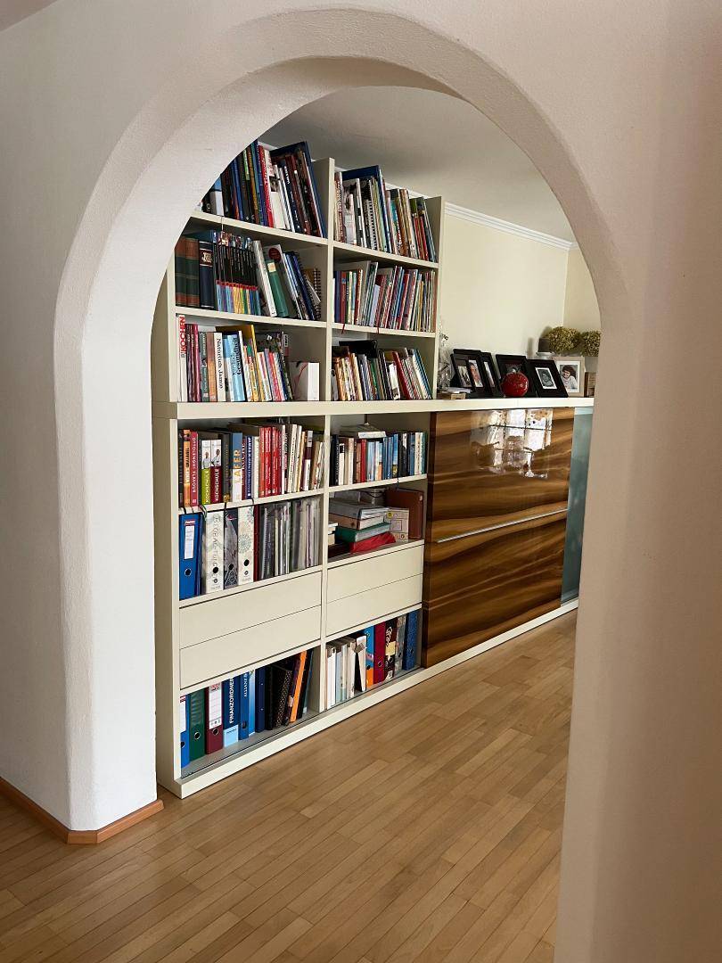Bücherregal in einem hellen Raum mit Holzboden und Rundbogen-Durchgang.