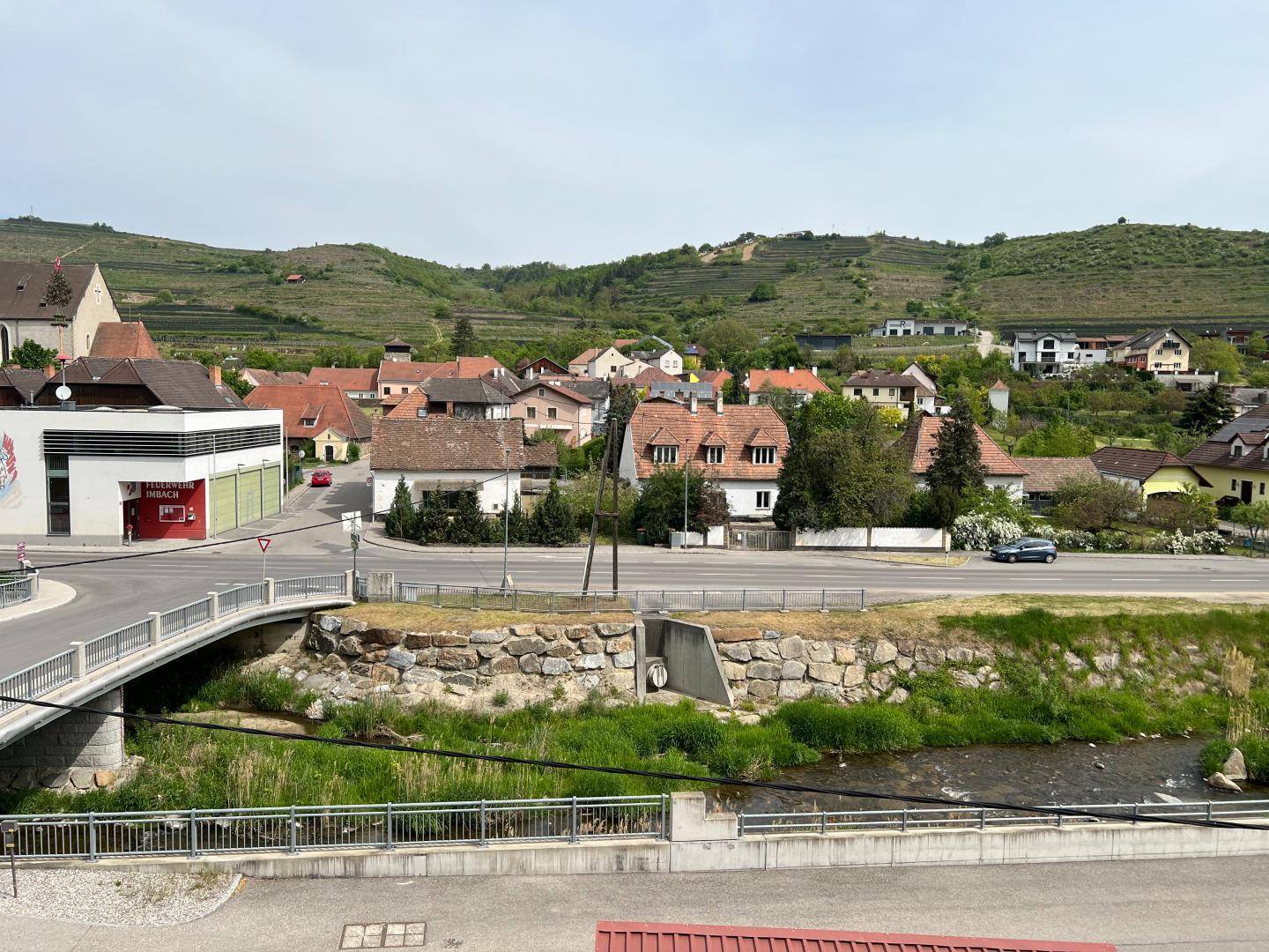 Weitblick über ein malerisches Dorf mit Fluss, Brücke, Weinbergen und grünen Hügeln im Hintergrund.