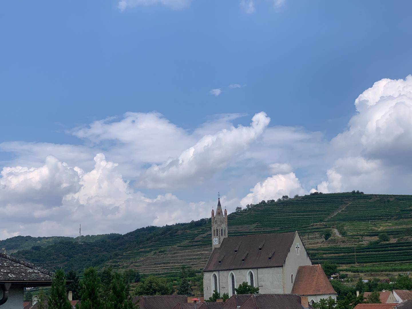 Malerischer Ausblick auf eine Kirche, ein Dorf, grüne Hügel und Weinberge unter blauem Himmel.