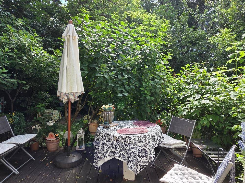 Sonnige Terrasse mit Holzboden, Gartentisch und Stühlen, umgeben von üppigem Grün.