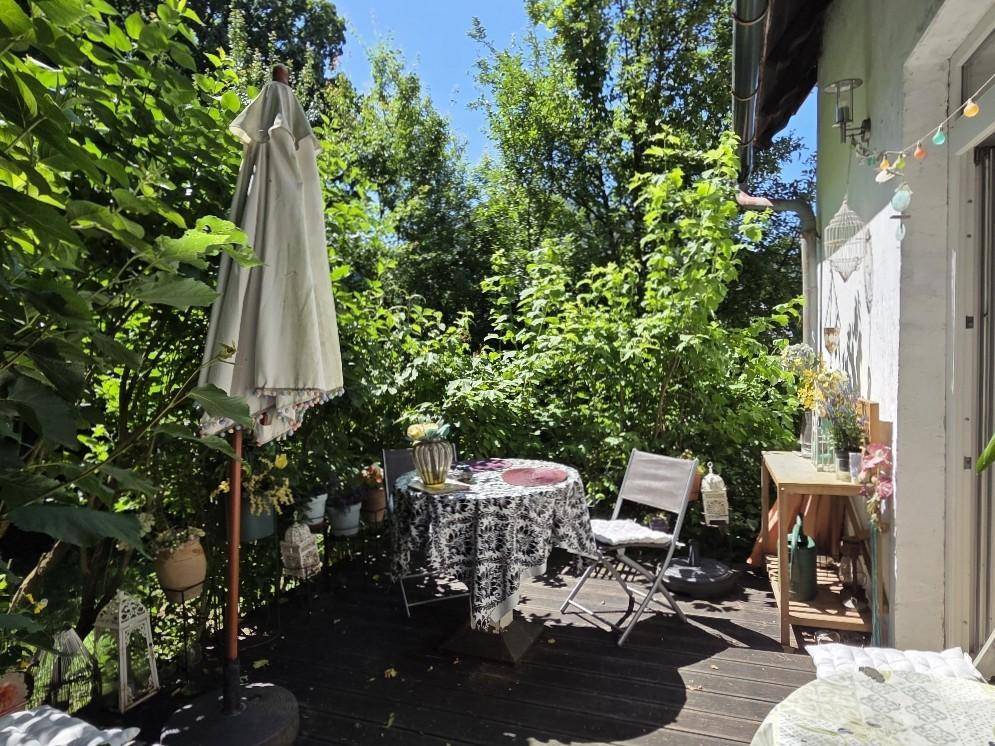 Sonnige Holzterrasse mit Gartentisch und Stühlen, umgeben von üppigem Grün.