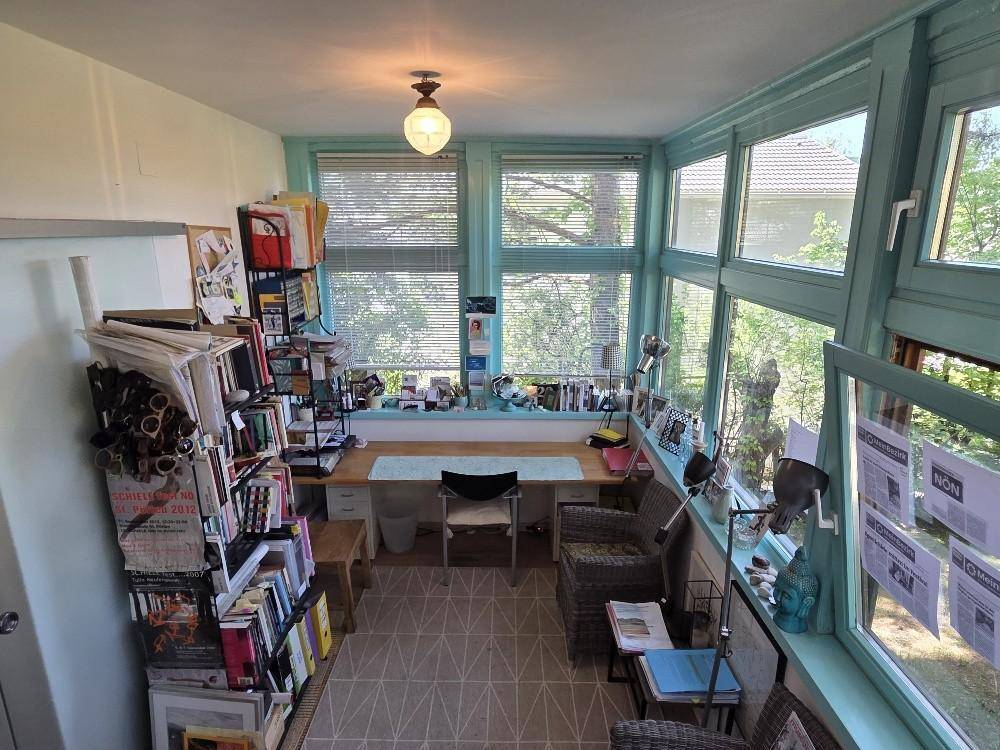 Helles Büro mit vielen Fenstern, Schreibtisch und Bücherregalen, umgeben von Grün.