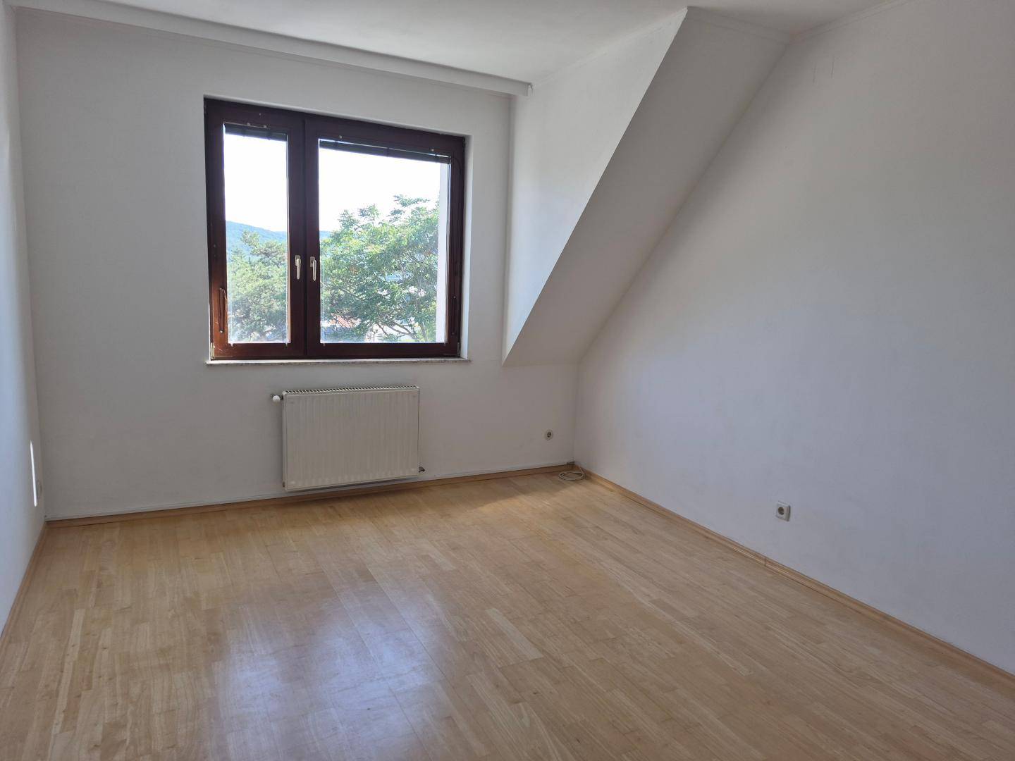 Schlafzimmer mit Laminatboden, Dachschräge und großem Fenster, das viel Tageslicht hereinlässt.