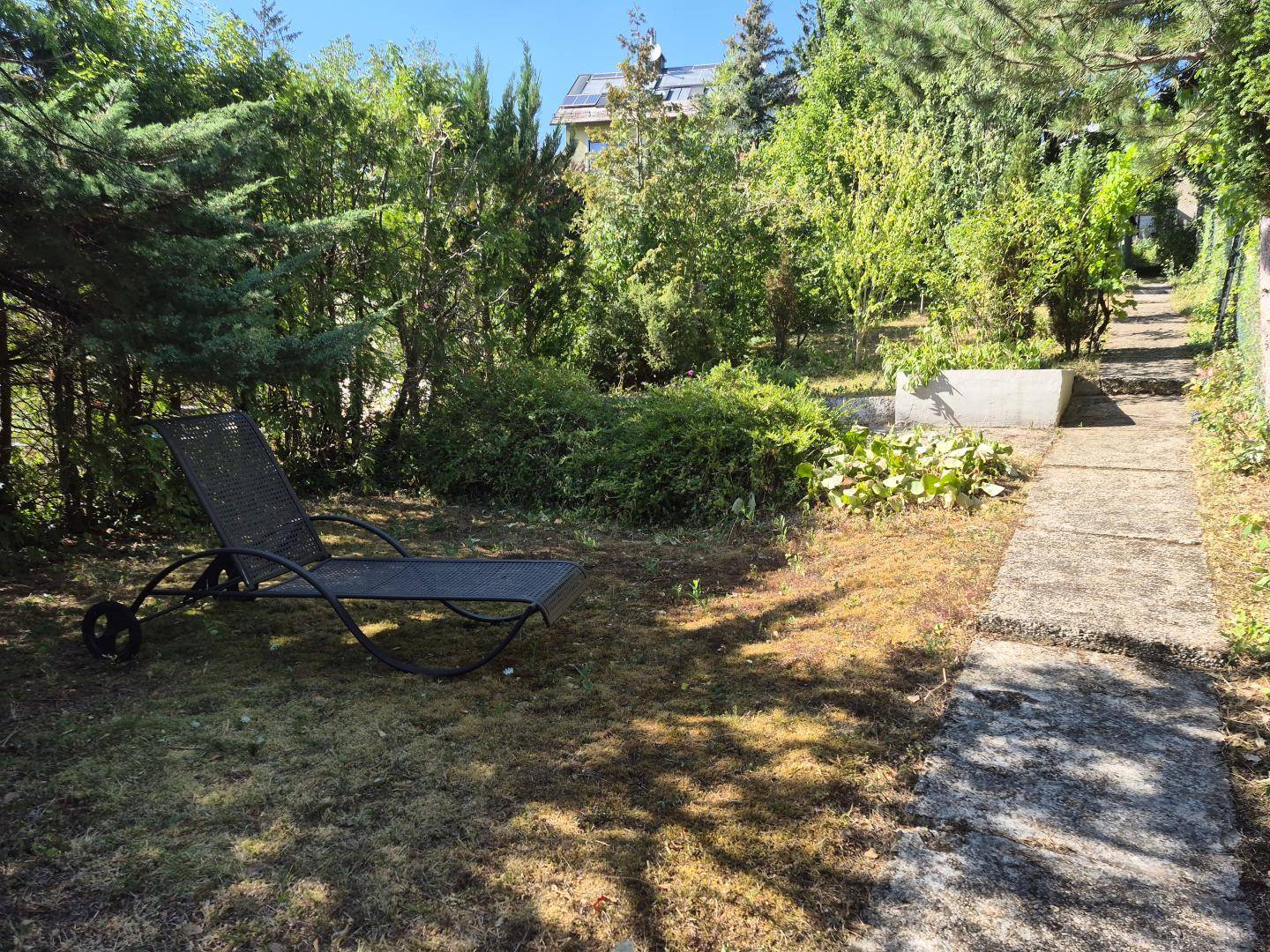 Gartenbereich mit Liegestuhl, gepflastertem Weg und viel Grün in sonniger Umgebung.