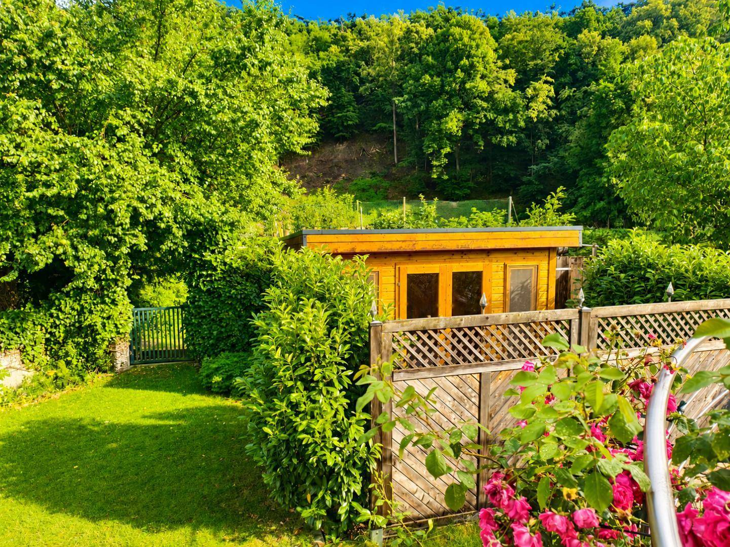 Gepflegter Garten mit Rasenfläche, Holzzaun und einem kleinen Gartenhaus umgeben von Bäumen.