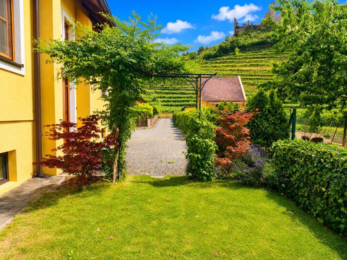 Gepflegter Gartenbereich mit Rasen und üppiger Vegetation, Blick auf Weinberge und Burgruine.