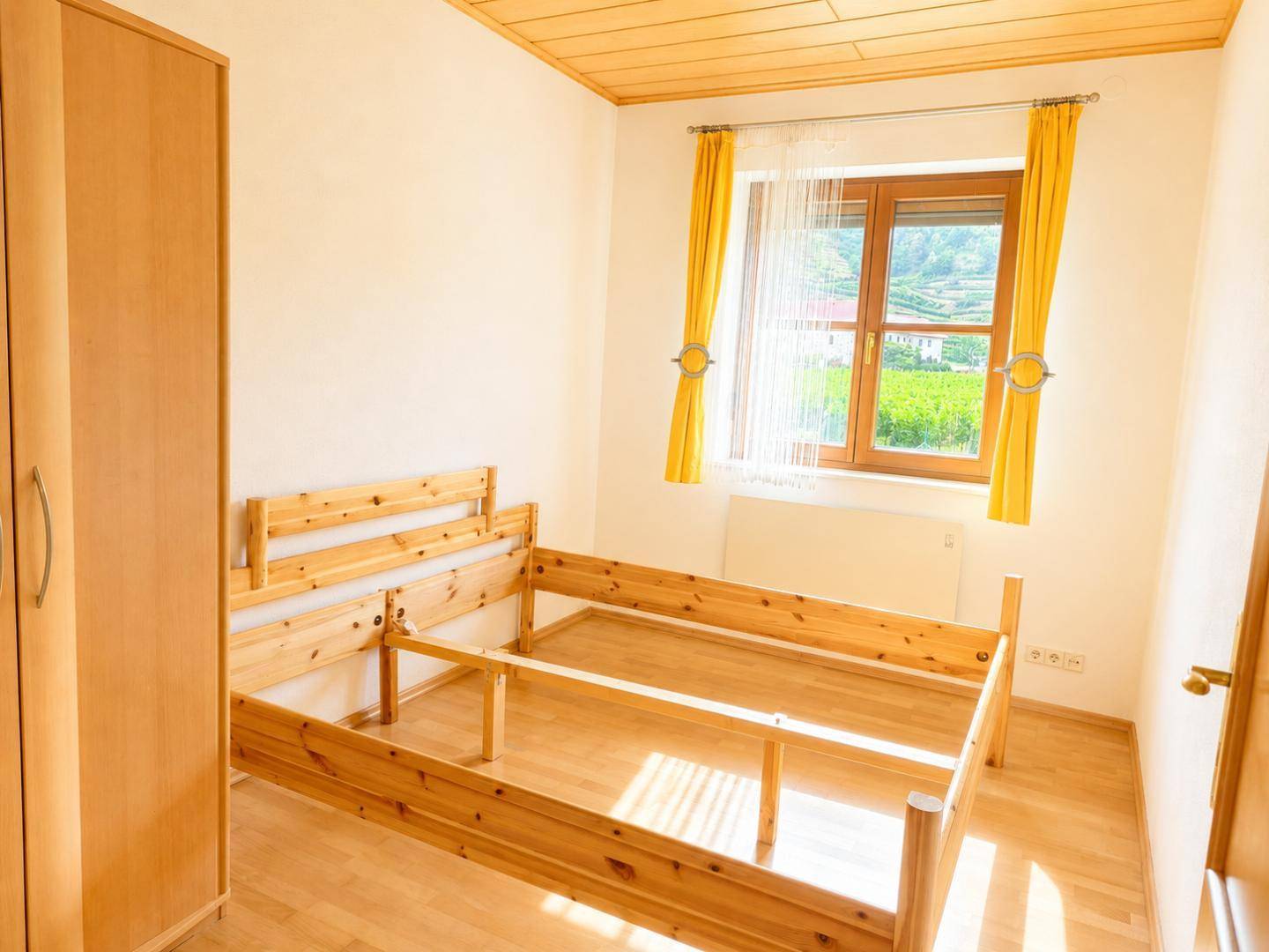 Helles Schlafzimmer mit Holzbettgestell, Kleiderschrank und Blick ins Grüne durch das Fenster.