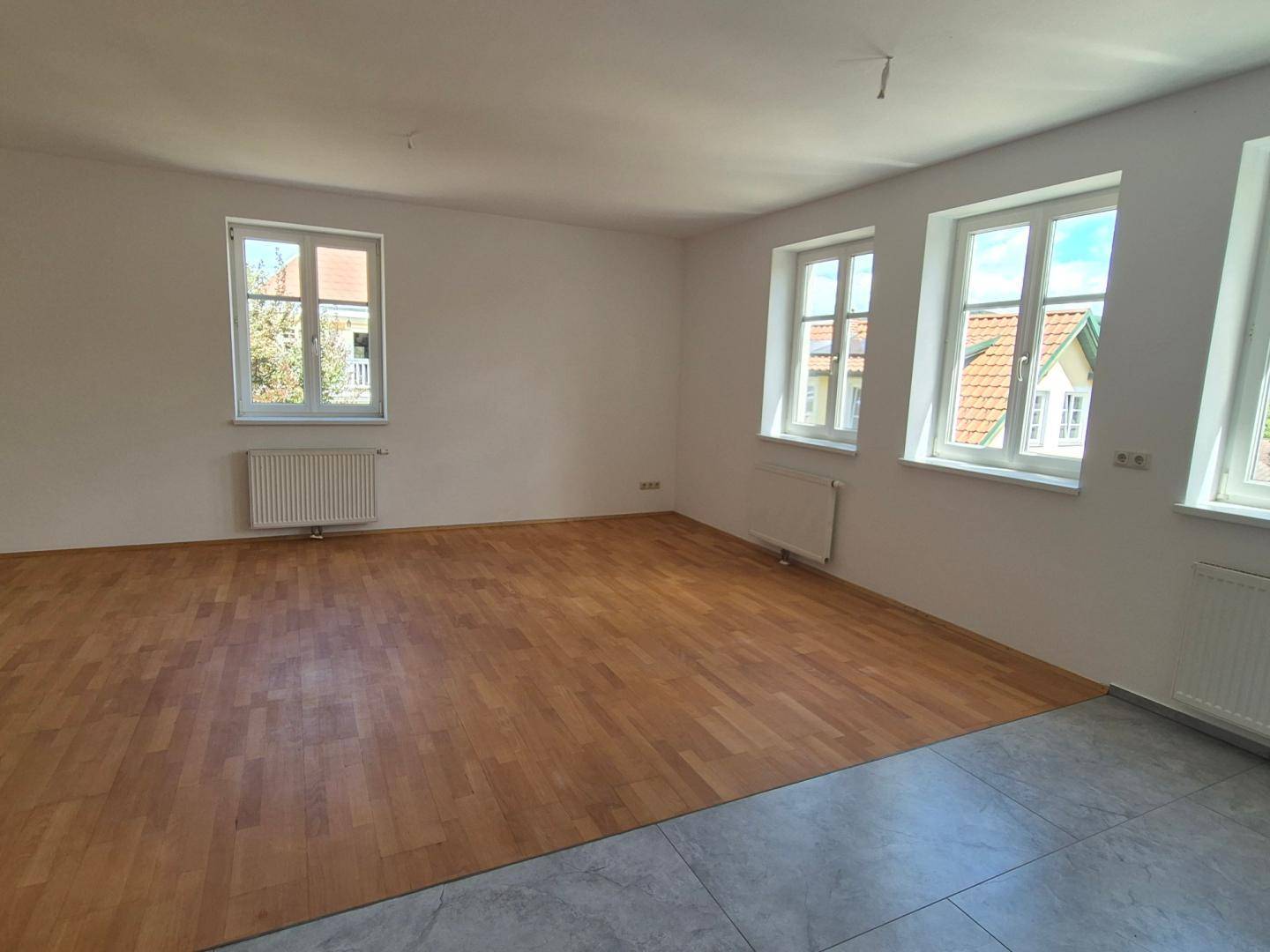 Heller Raum mit gemischtem Bodenbelag aus Holz und Fliesen sowie mehreren Fenstern.