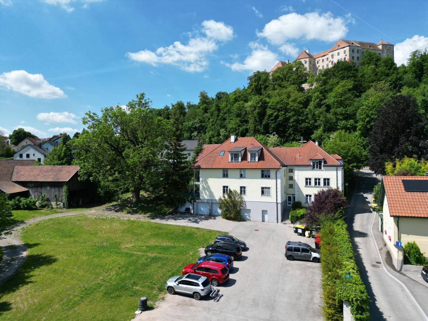 Luftaufnahme des Mehrfamilienhauses mit Parkplätzen und ländlicher Umgebung, Burg im Hintergrund.