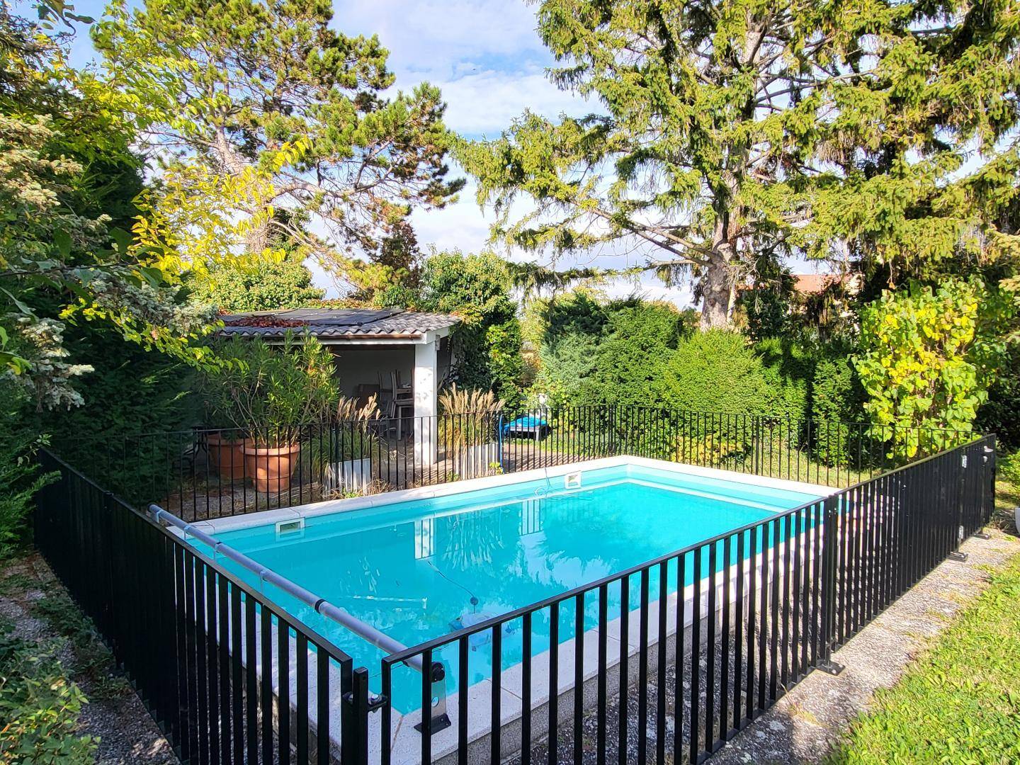 Eingezäunter Swimmingpool im Garten, umgeben von Bäumen und Sträuchern, mit einem kleinen überdachten Bereich im Hintergrund.