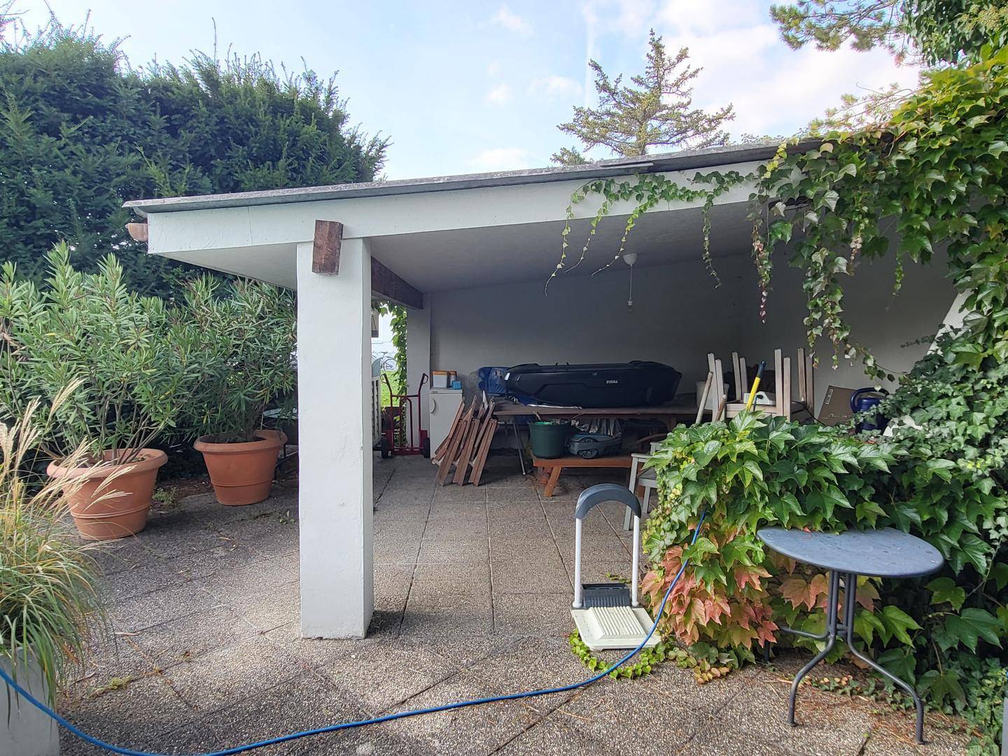 Überdachter Außenbereich mit gepflastertem Boden, umgeben von Pflanzen, der als Abstellfläche oder Terrasse genutzt wird.