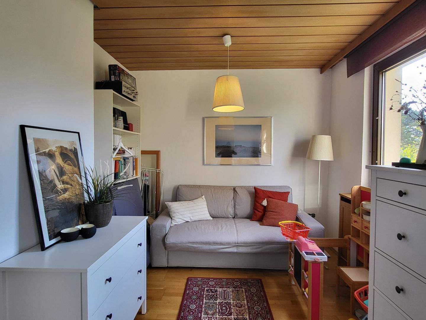 Gemütliches Wohnzimmer mit hellem Sofa, Holzdecke und Blick ins Grüne durch das Fenster.