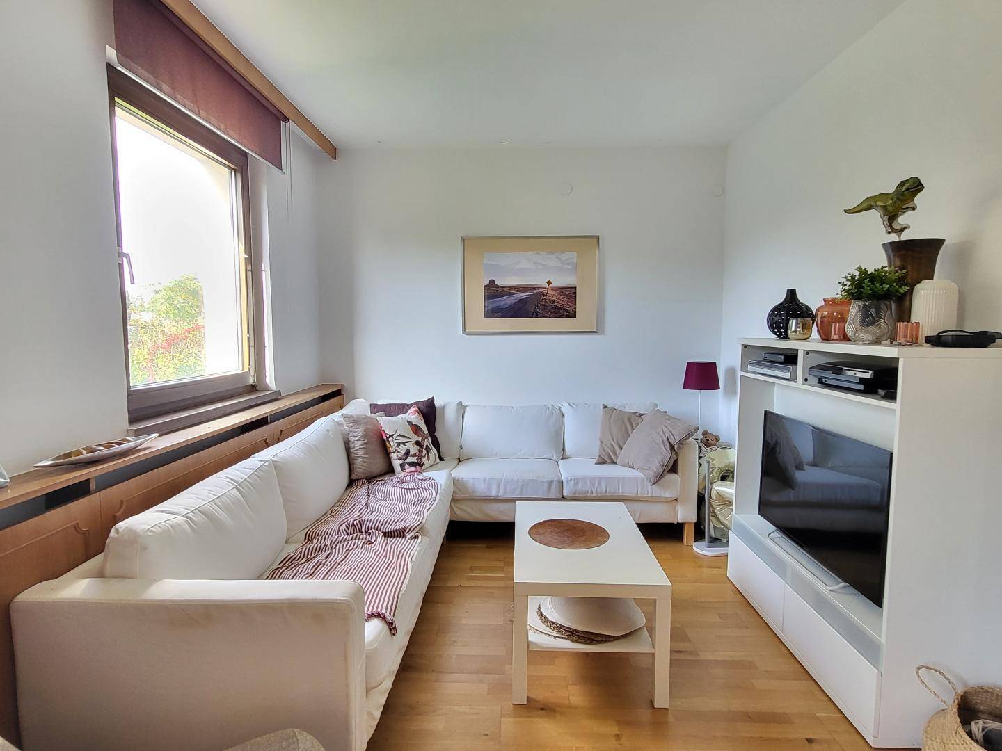 Helles Wohnzimmer mit großem Ecksofa, Holzboden und einem Fenster mit Blick nach draußen.
