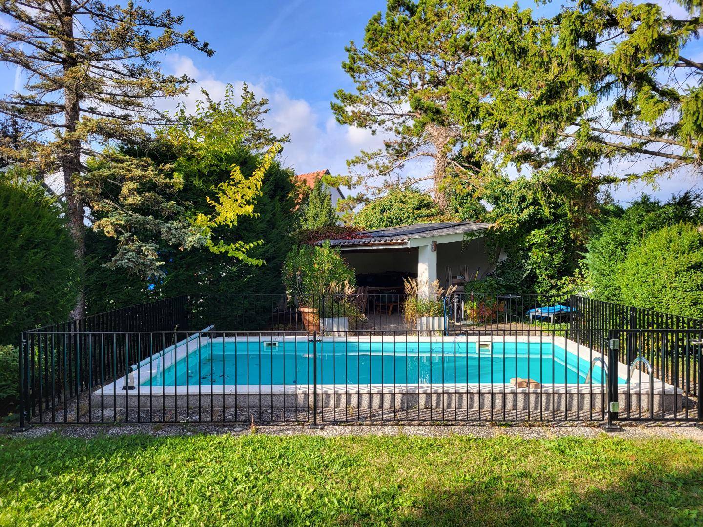 Umzäunter Swimmingpool im Garten mit angrenzendem überdachtem Bereich und viel Grün.