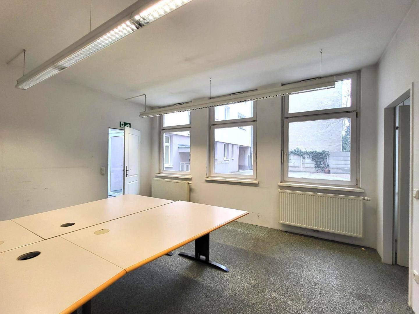 Leerer Büroraum mit großem Schreibtisch und drei Fenstern mit Blick auf den Innenhof.