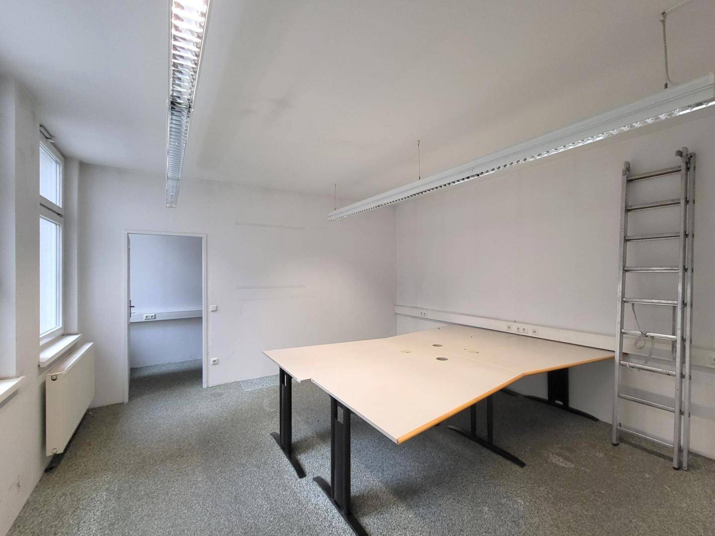 Leerer Büroraum mit großem Schreibtisch, einer Leiter und Fenstern, die Tageslicht hereinlassen.