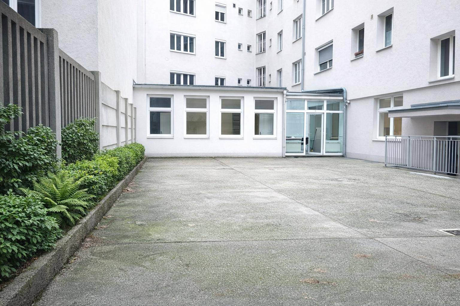 Leerer Außenparkplatz mit Betonboden, umgeben von einem Gebäude und einer gepflegten Hecke.