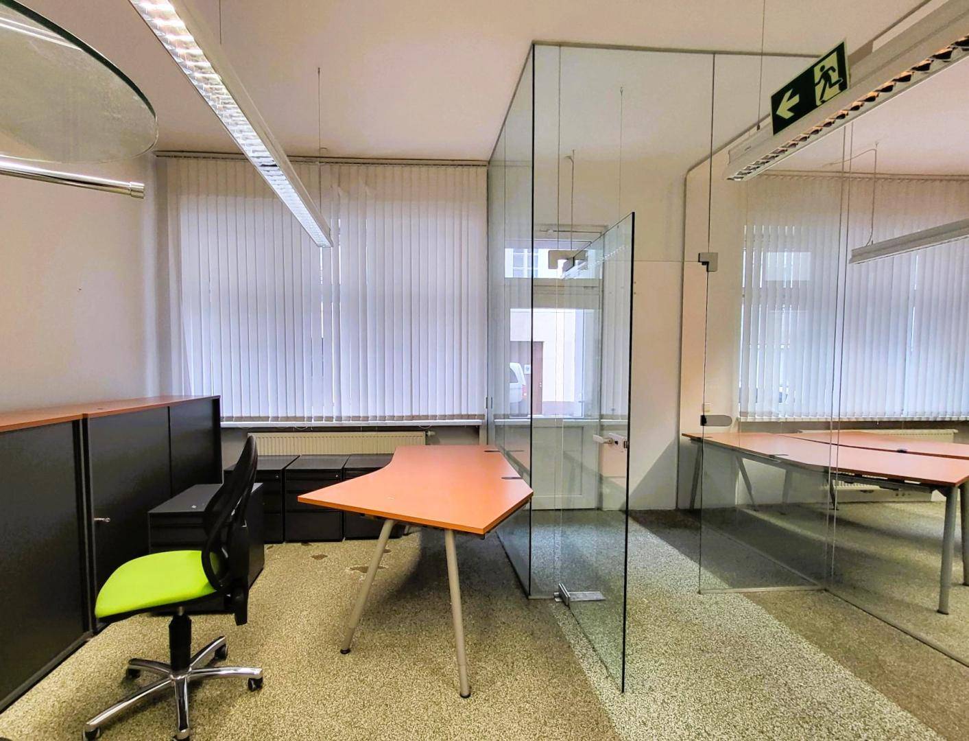 Büro mit Glaswänden, Schreibtischen und einem grünen Bürostuhl, Fenster mit Jalousien für Privatsphäre.