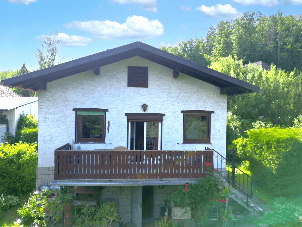 Vorderansicht des Hauses mit weißer Fassade, braunem Holzbalkon und großen Fenstern.