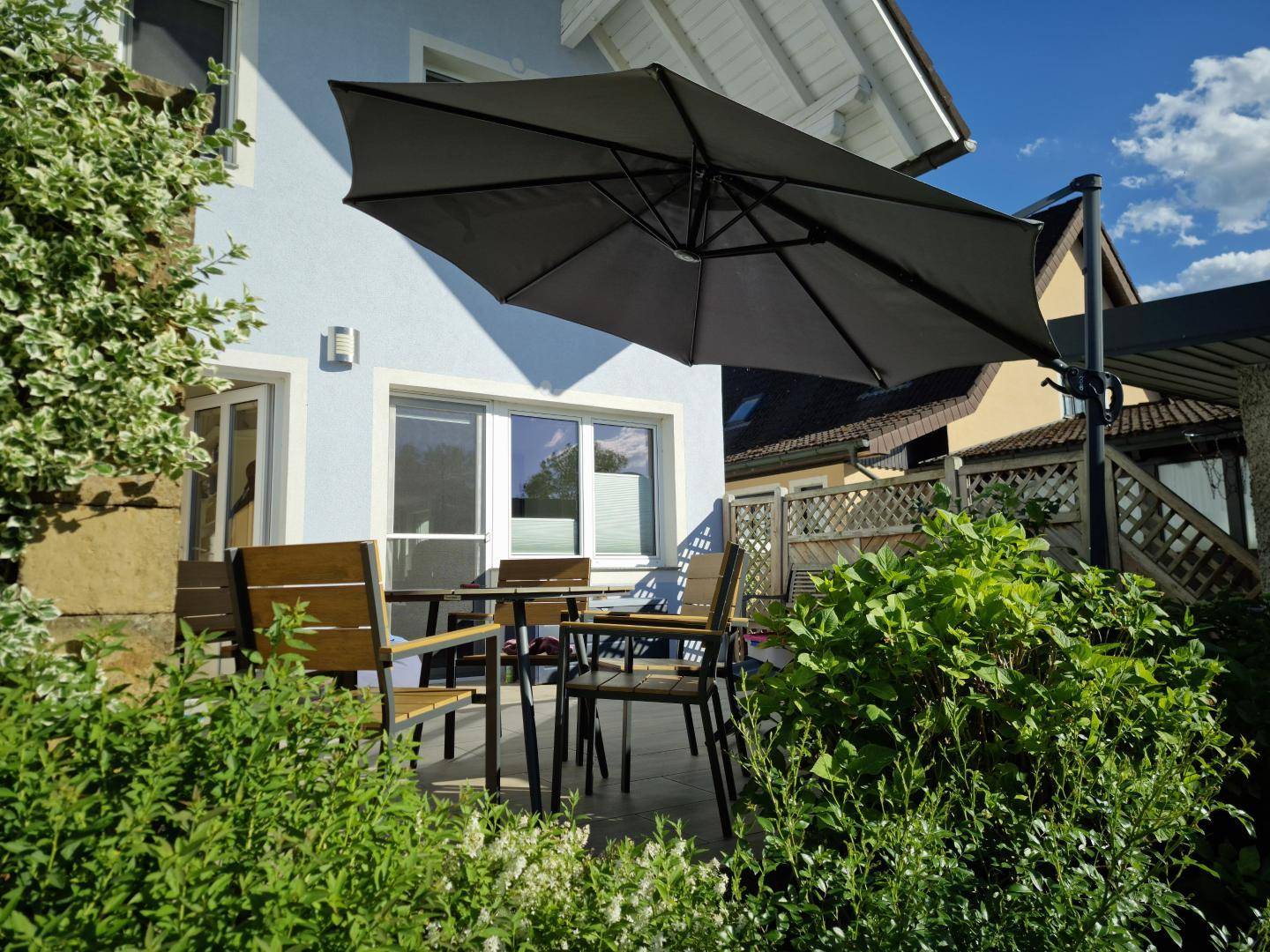 Sonnige Terrasse direkt am Haus mit Gartenmöbeln und großem Sonnenschirm, umgeben von grünen Büschen.