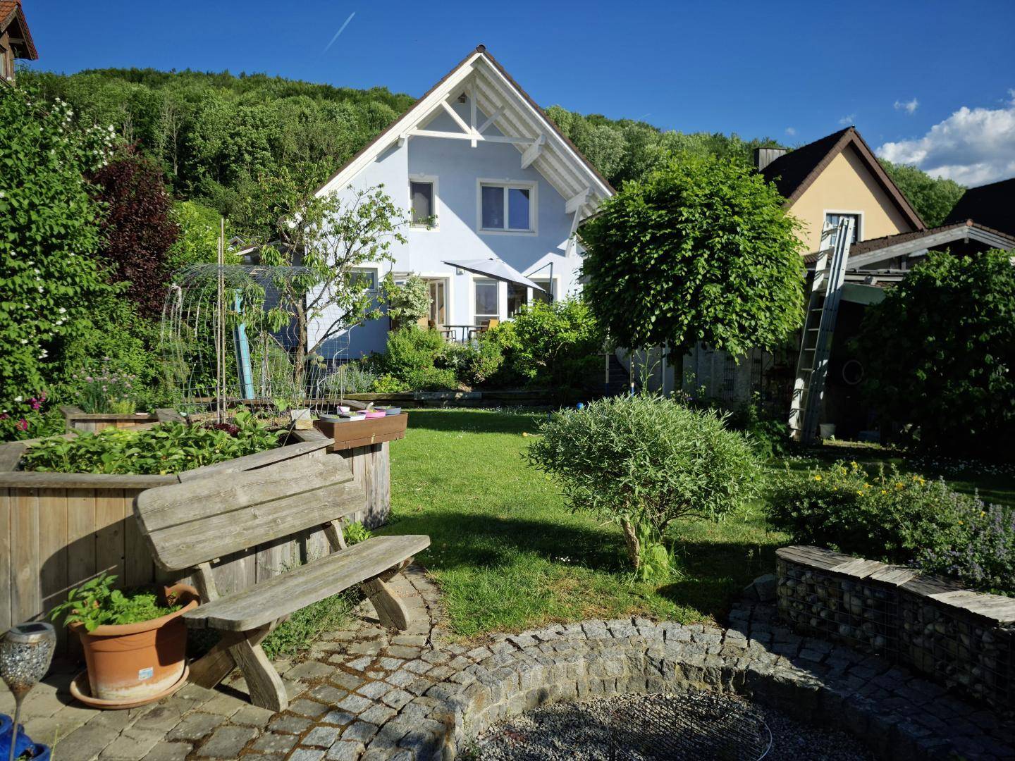 Gepflegter Garten mit Rasenfläche, Holzbank und einer Feuerstelle, umgeben von grünen Pflanzen.