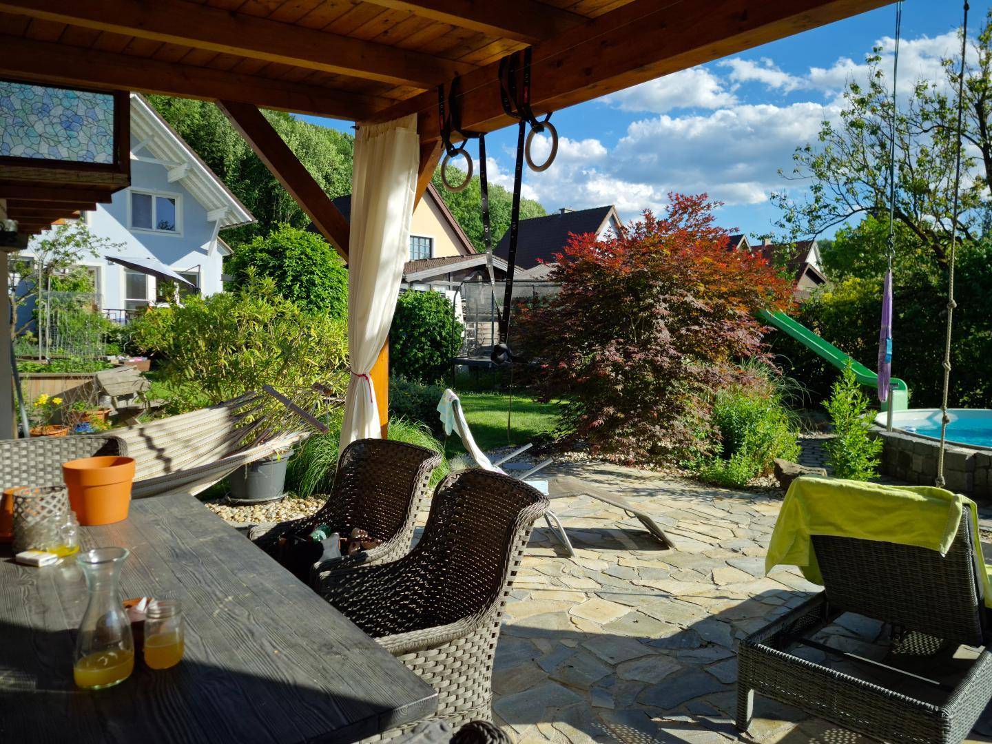 Überdachte Terrasse mit Rattanmöbeln und Blick auf den Garten mit Pool und Spielgeräten.