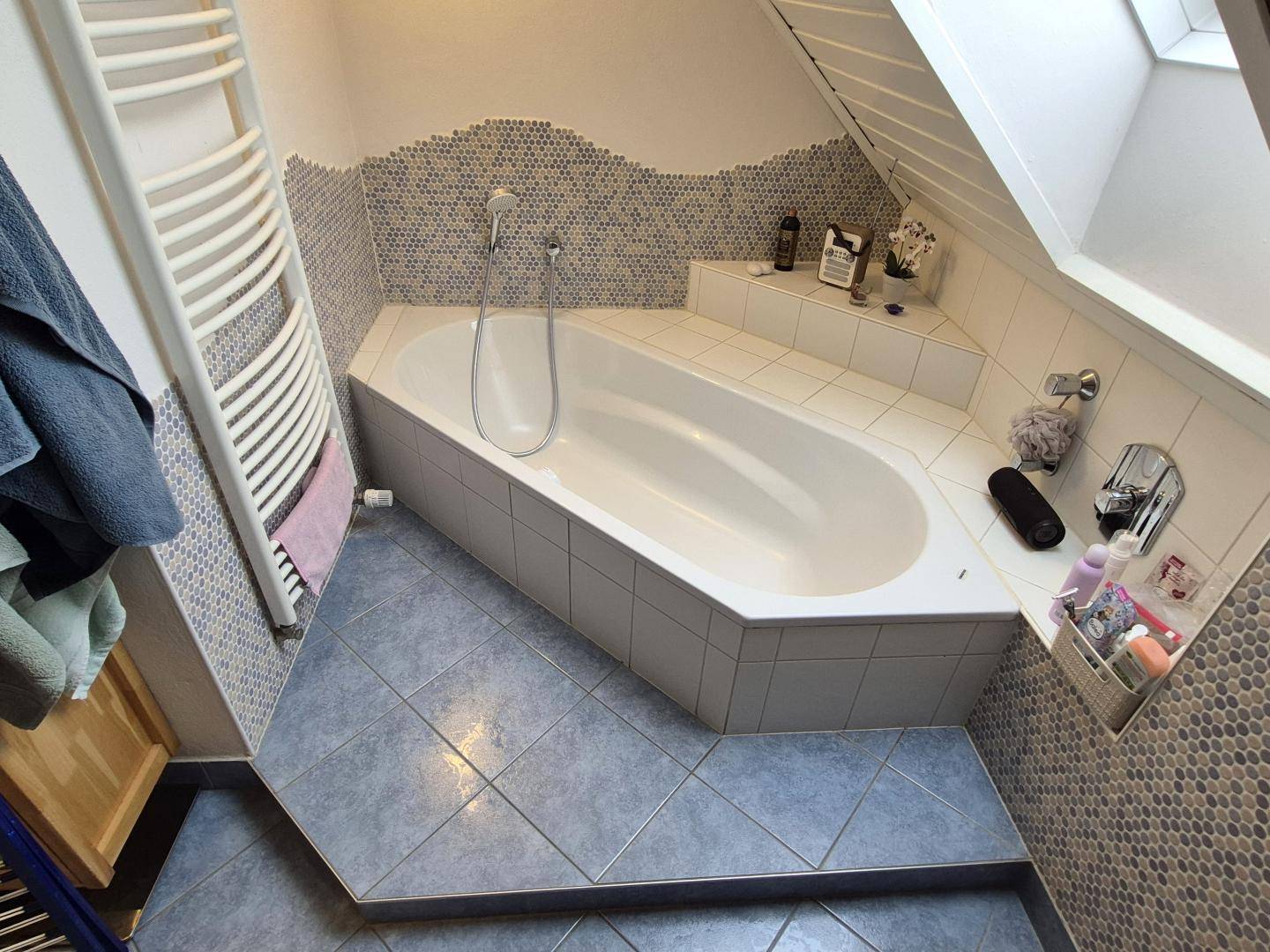 Badezimmer mit Eckbadewanne, blauen Fliesen und praktischem Handtuchheizkörper unter dem Dachfenster.
