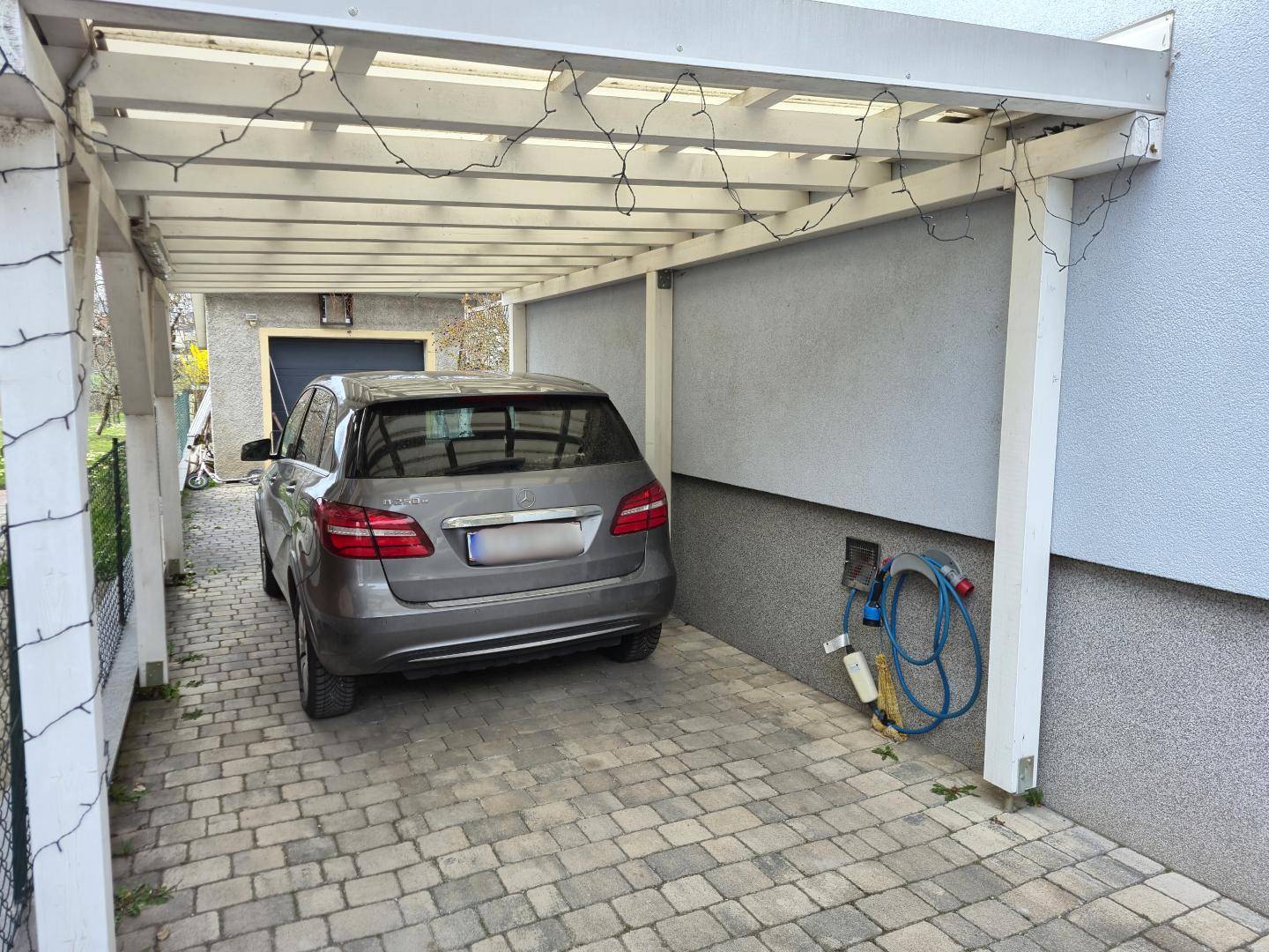 Überdachter Carport mit gepflastertem Stellplatz und Ladestation für Elektrofahrzeuge.