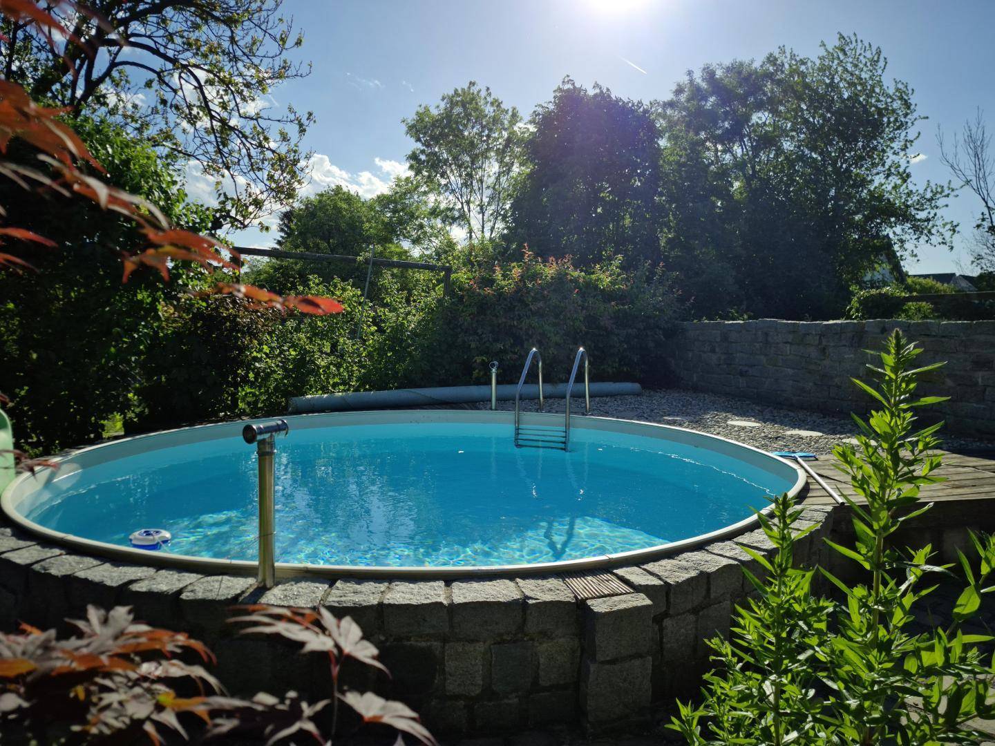 Runder Aufstellpool im Garten, umgeben von Steinmauern und üppigem Grün unter strahlend blauem Himmel.