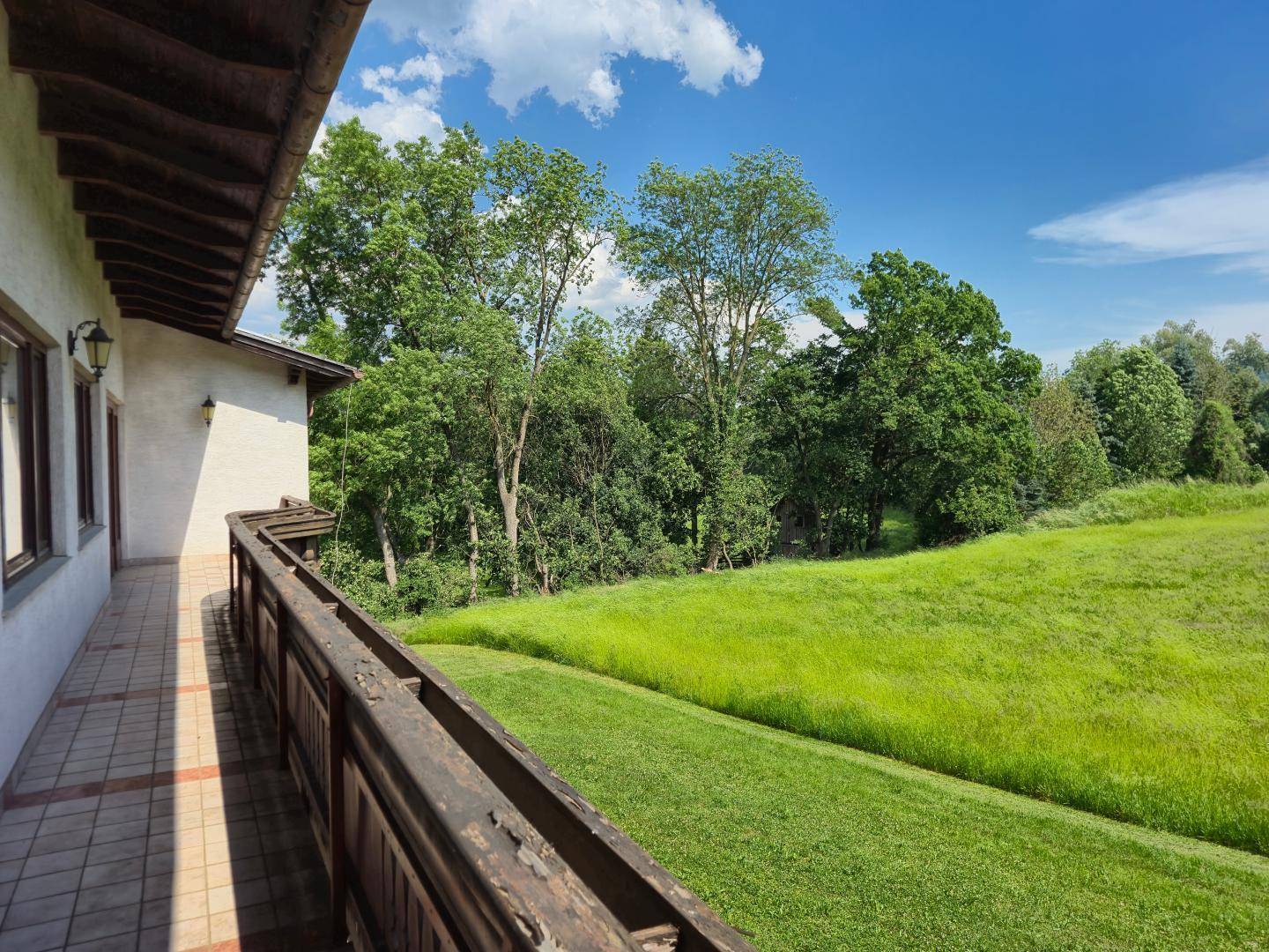 Langer Balkon mit Holzbalken und Fliesenboden, bietet einen weiten Blick auf grüne Wiesen und Bäume.