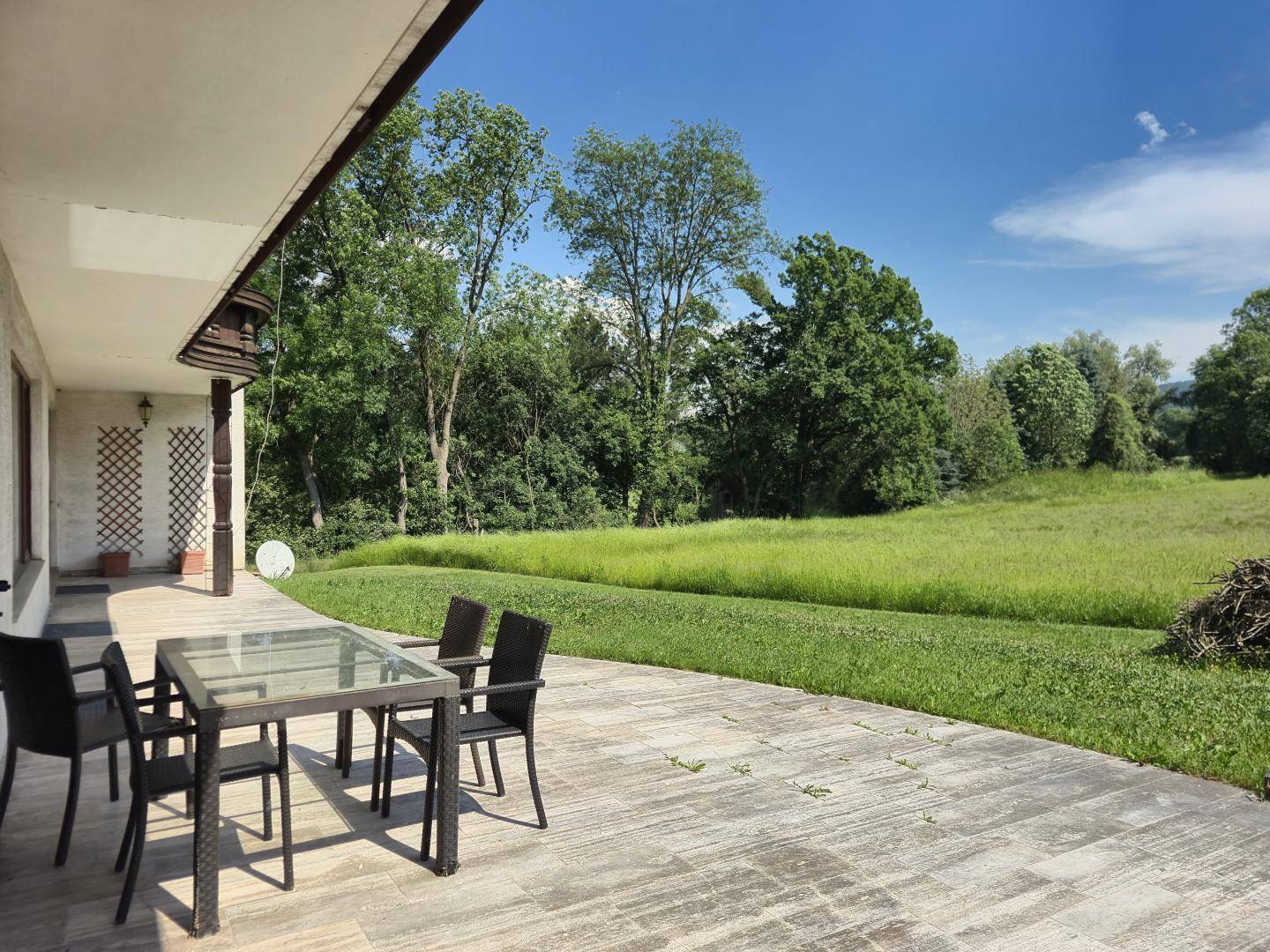 Große Terrasse mit Esstisch und Stühlen, umgeben von grünen Wiesen und Bäumen.