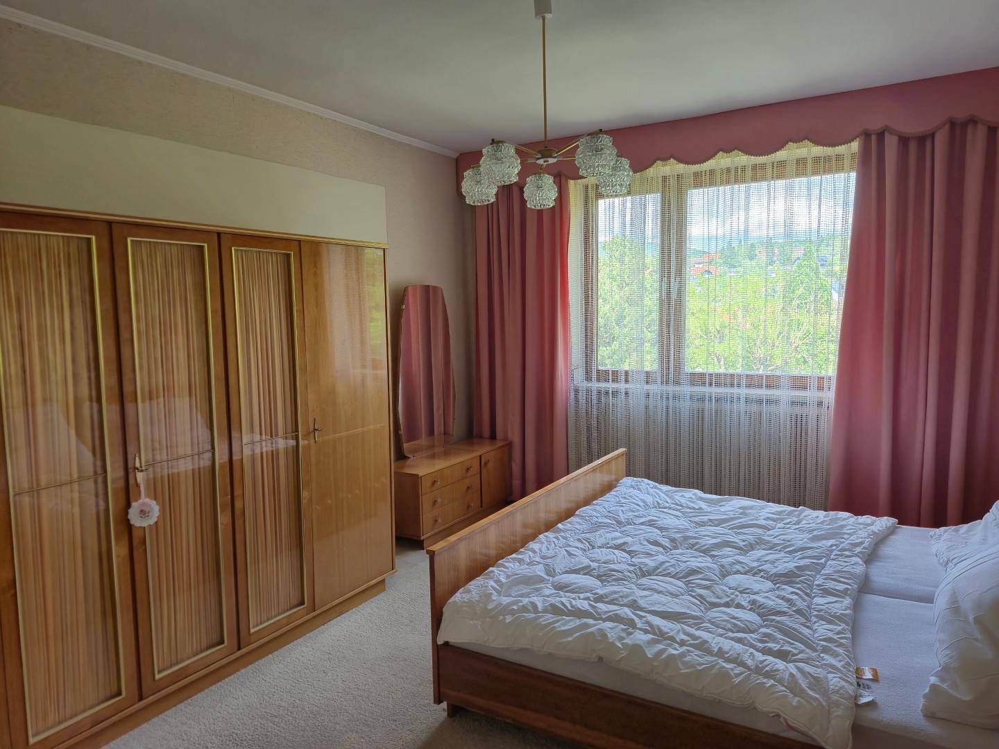 Gemütliches Schlafzimmer mit Holzmöbeln, Teppichboden und rosa Vorhängen, mit Blick ins Grüne.