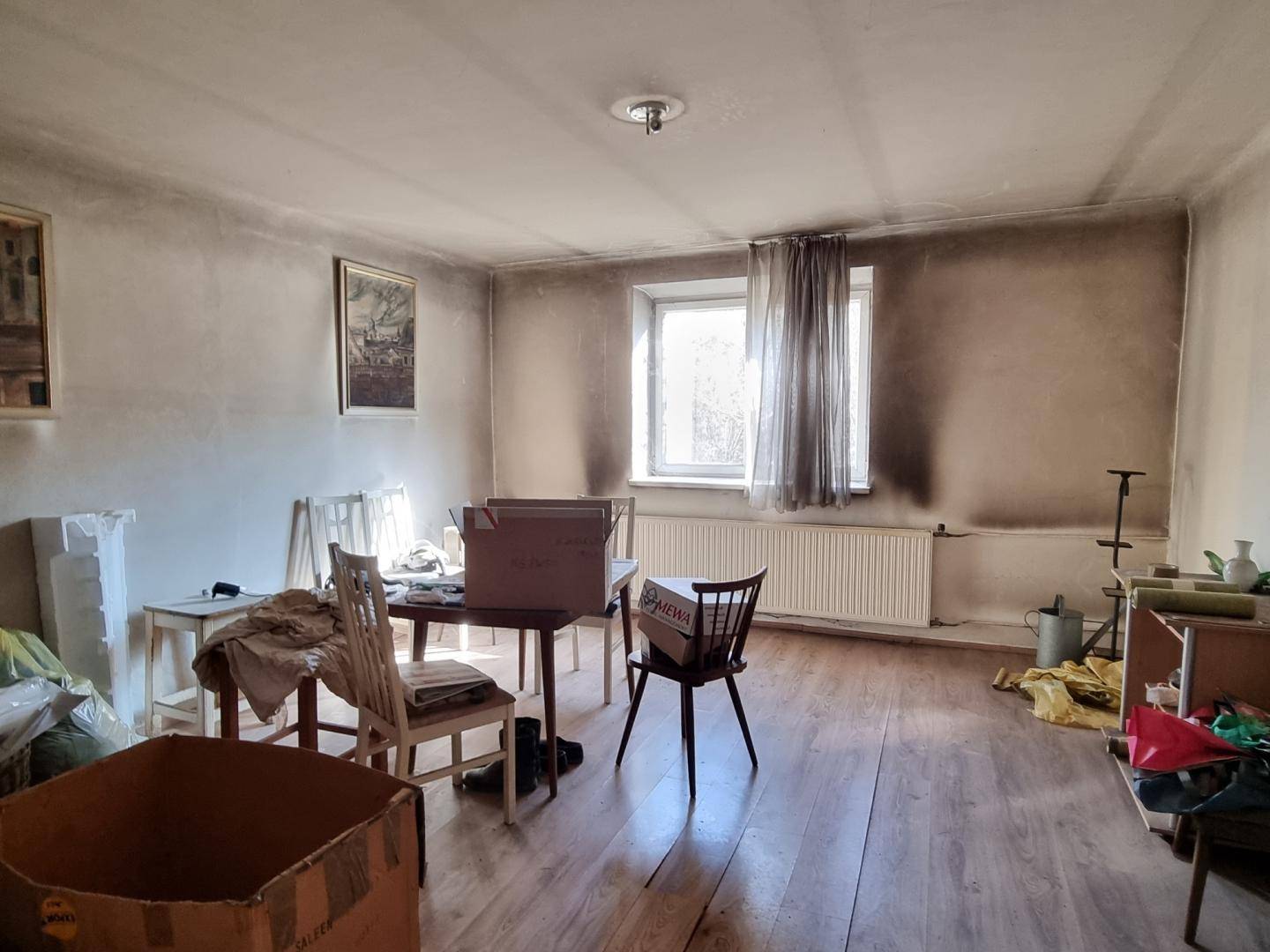 Wohnraum mit Holzboden, Fenster und Spuren von Renovierungsbedarf.