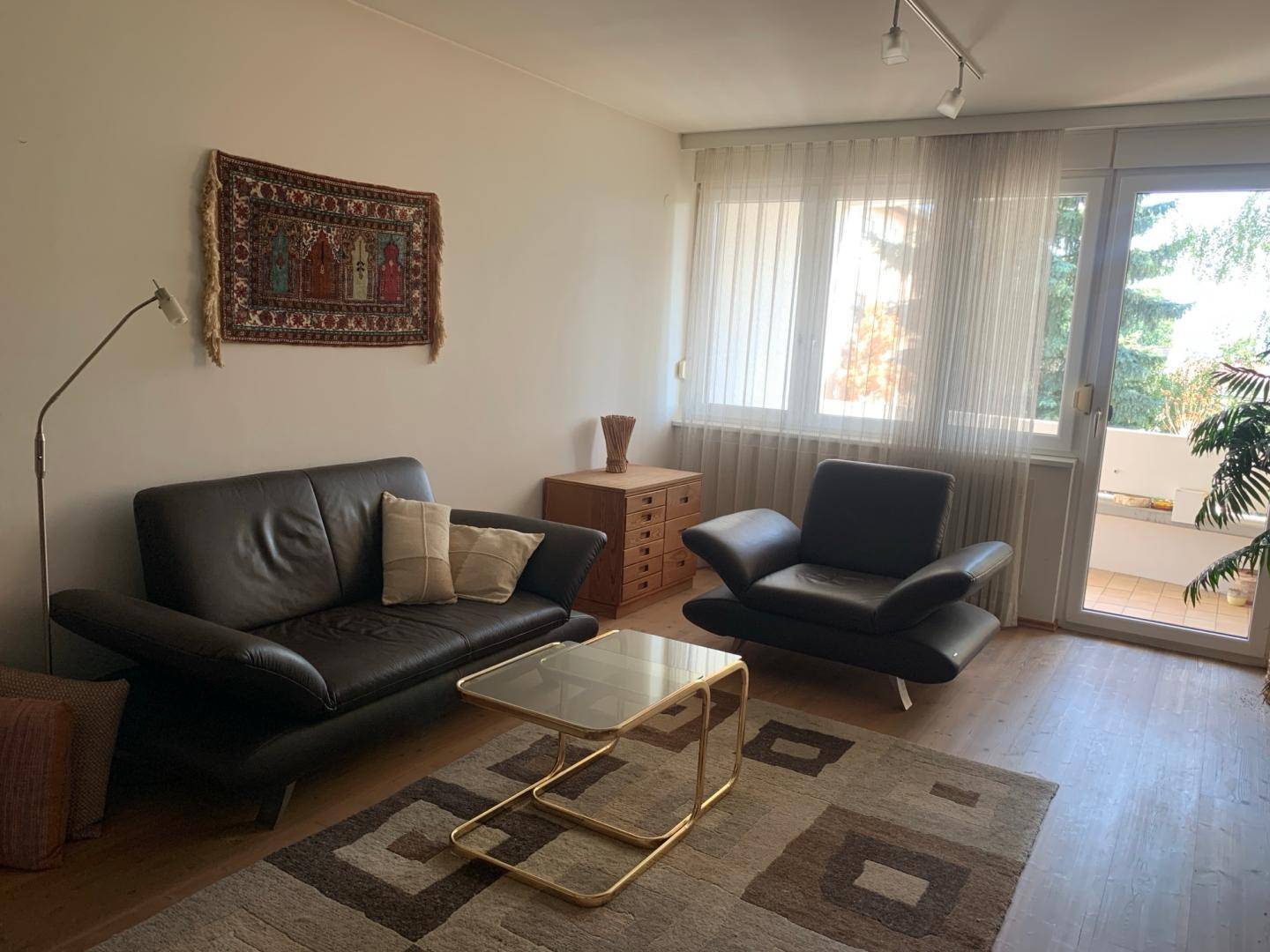 Helles Wohnzimmer mit Zugang zum Balkon, dunklen Ledermöbeln und einem Teppich.