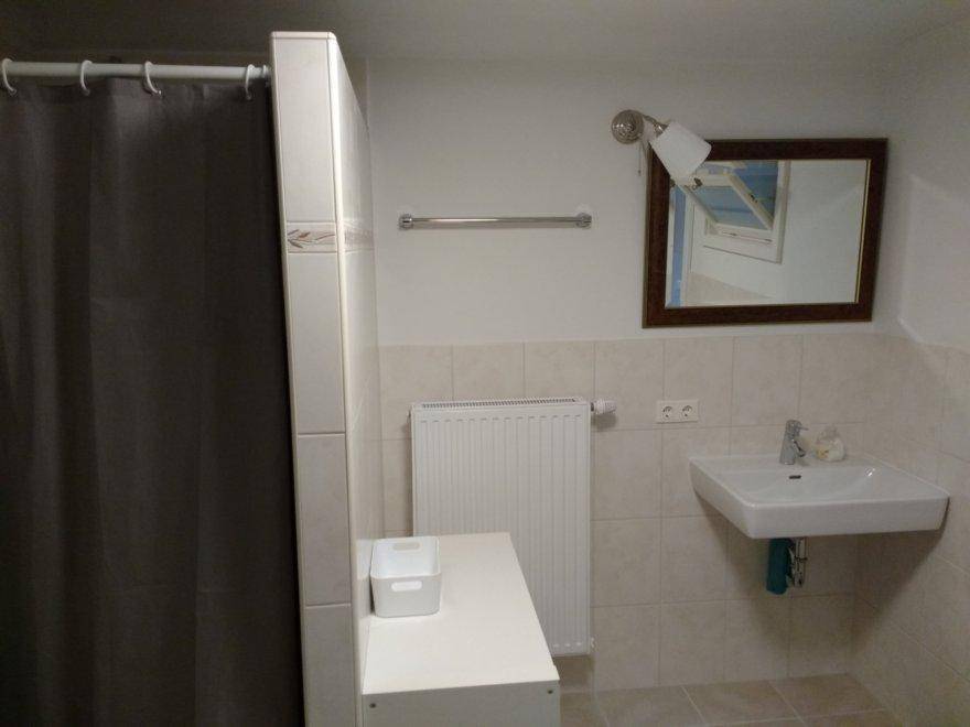 Badezimmer mit hellen Fliesen, wandmontiertem Waschbecken, Spiegel und Duschbereich.