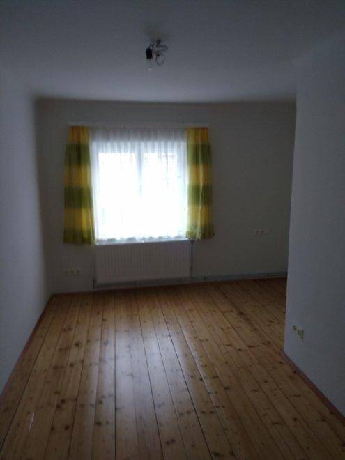 Leeres Zimmer mit Holzdielenboden und einem Fenster mit gestreiften Vorhängen.