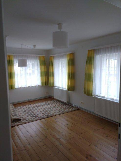 Helles Zimmer mit Holzdielenboden und drei Fenstern mit farbigen Vorhängen.