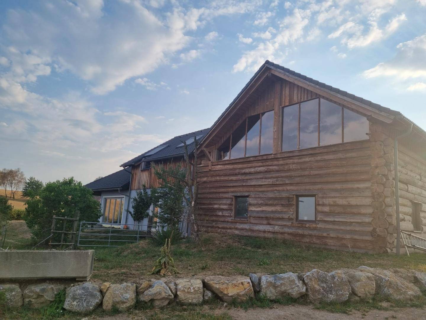 Rustikales Holzblockhaus mit großen Giebelfenstern in ländlicher Umgebung unter blauem Himmel.