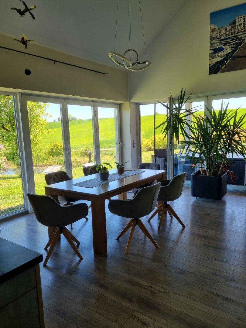Heller Essbereich mit Holztisch und modernen Stühlen, umgeben von großen Fenstern mit Grünblick.