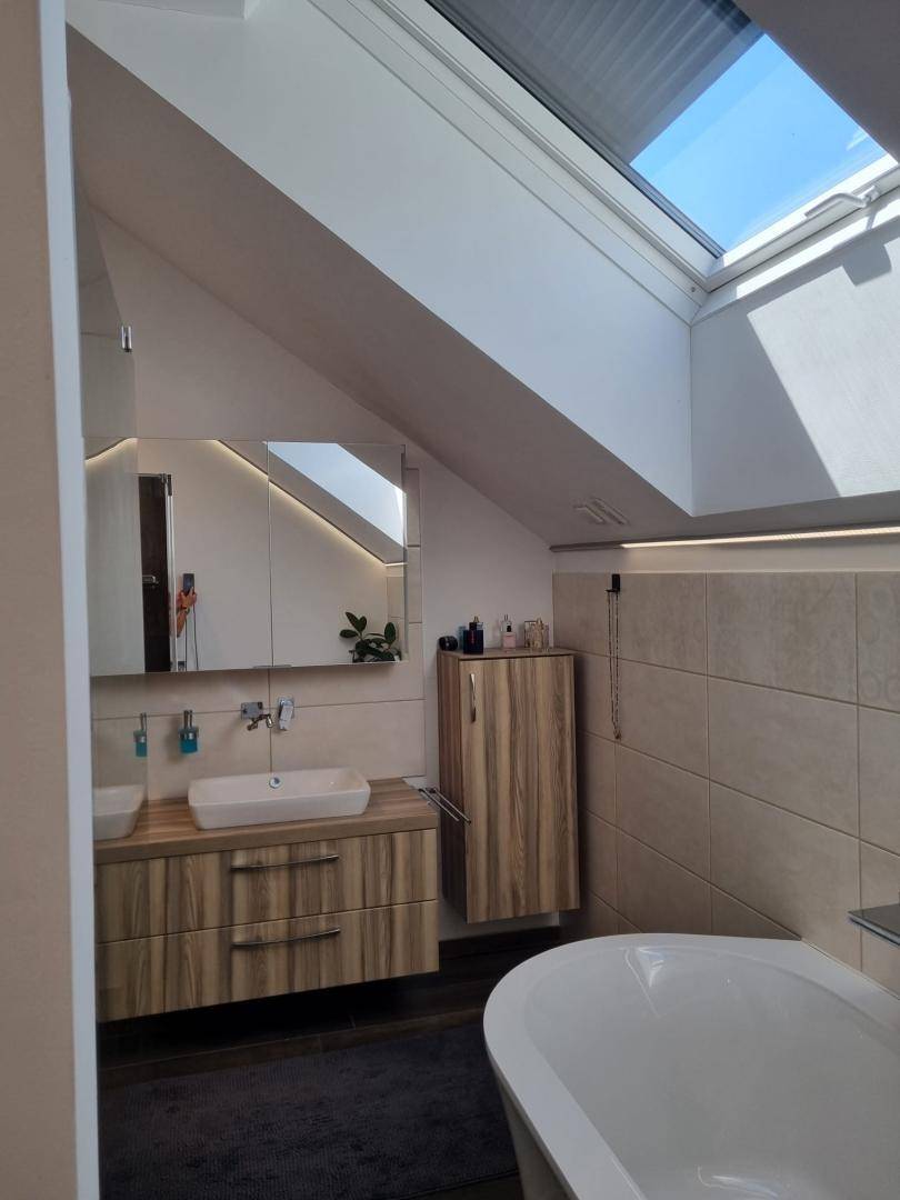 Heller Badezimmer-Waschtisch mit modernem Waschbecken, Spiegelschrank und Holzunterschränken unter einem Dachfenster.