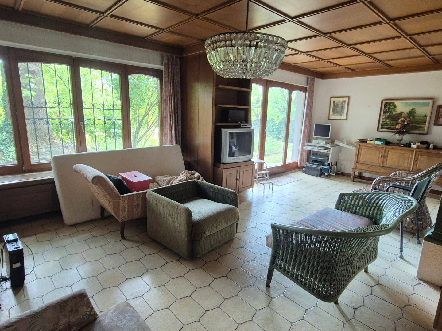 Geräumiges Wohnzimmer mit Holzkassettendecke, Fliesenboden und großen Fenstern zum Garten.